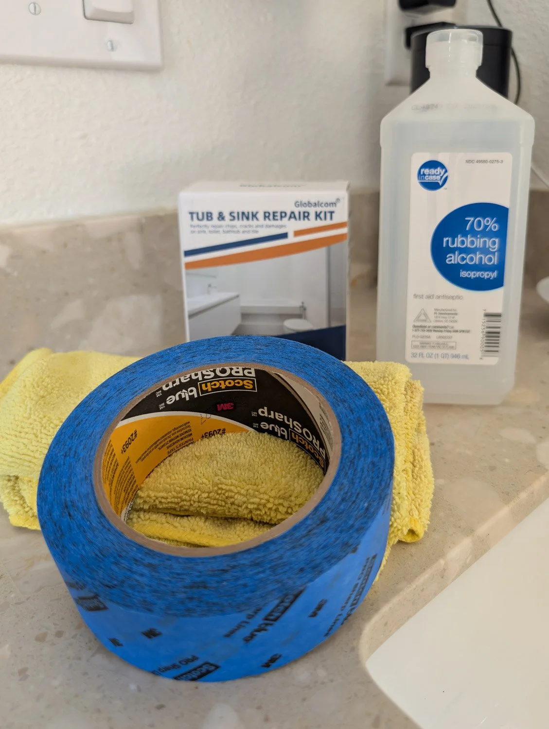 A collection of DIY repair supplies on a marble countertop, including a "Tub & Sink Repair Kit," a large bottle of rubbing alcohol, a roll of blue painter's tape, and a yellow microfiber cloth.