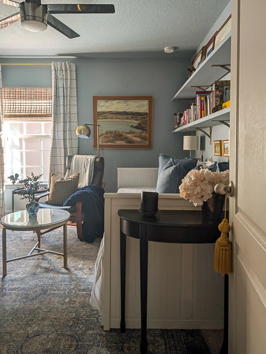 A cozy room features light blue walls, a white daybed with blue pillows, and open wall shelving filled with books and decor.