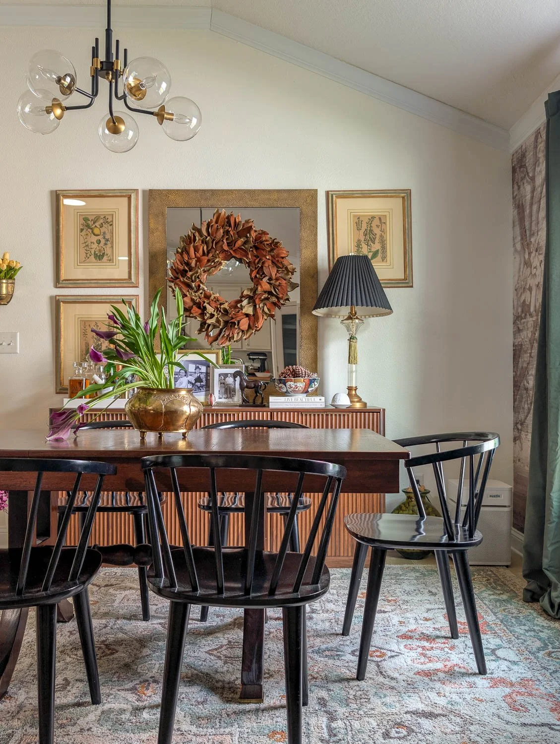 A traditional dining room featuring a wood table and black spindle chairs on a faded vintage-style rug, anchored by a fluted sideboard styled with a large gold-framed mirror, a magnolia wreath, and botanical art prints.