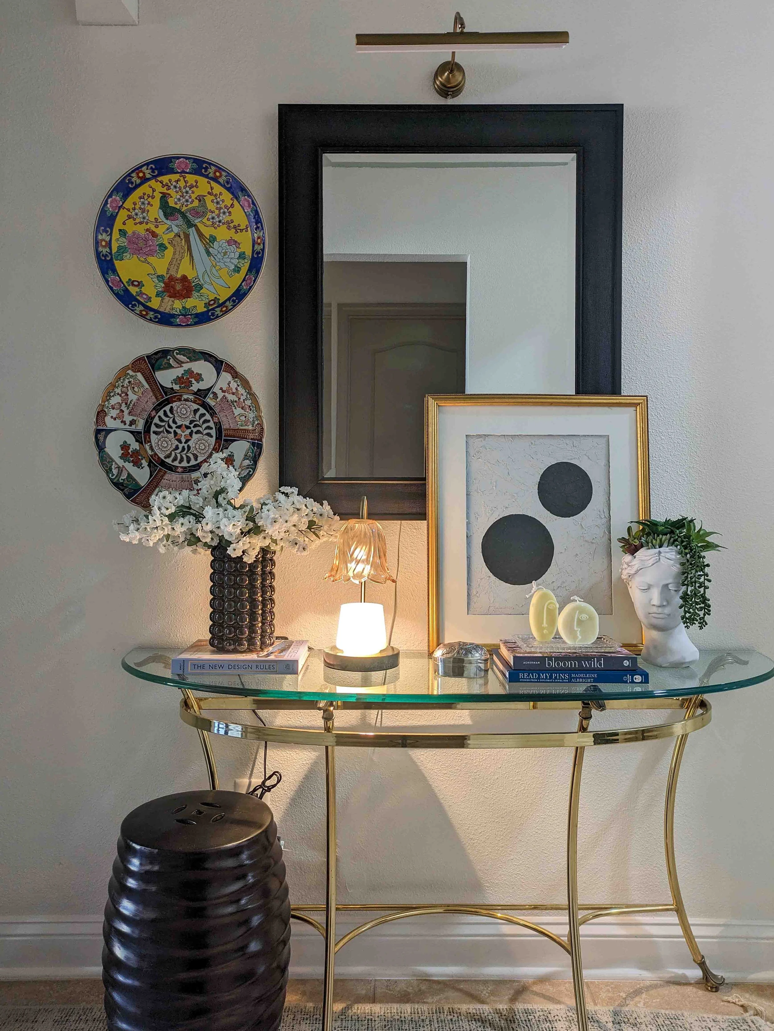 A brass and glass console table styled with a black textured vase of white flowers, intentional book stacks, a small glass lamp, and a bust planter against a white wall with a large black-framed mirror.