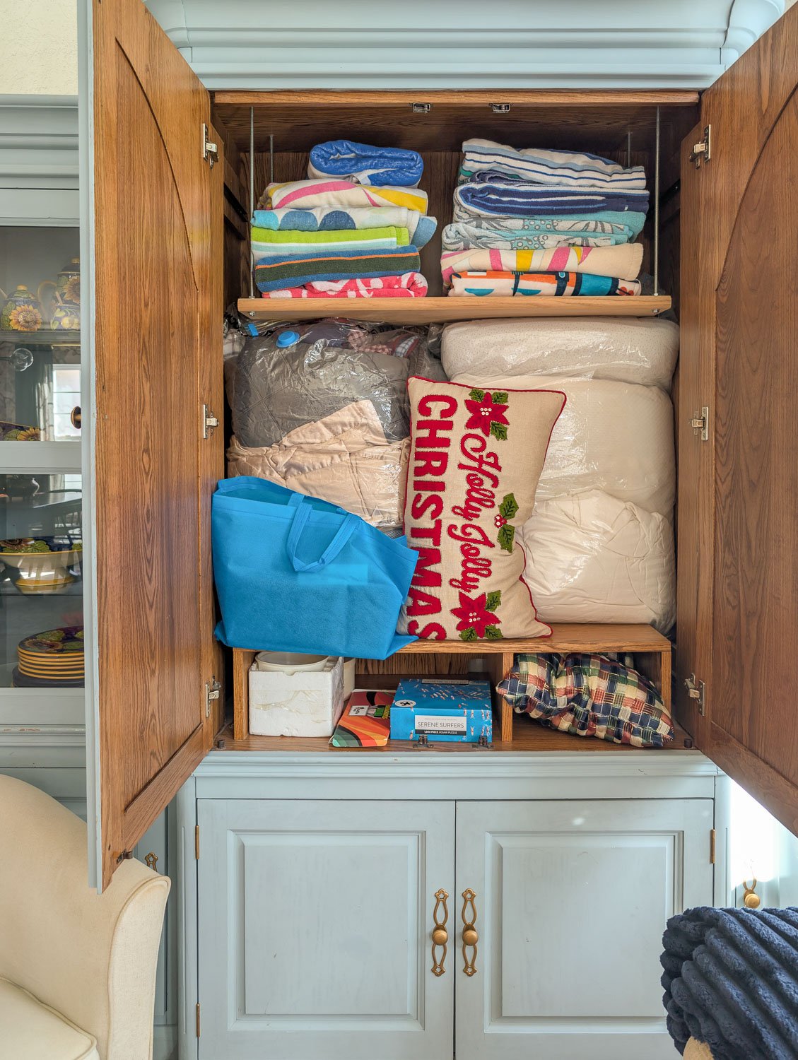 A 90s honey oak media cabinet with open doors filled to the brim with beach towels, blankets, and a Holly Jolly Christmas pillow.