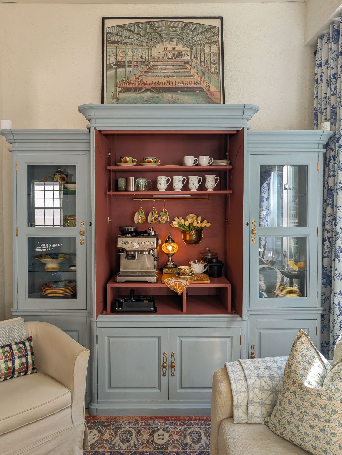 A wide shot of a large blue painted hutch repurposed as a coffee bar in a living room, styled with a rug, sofa, and art.