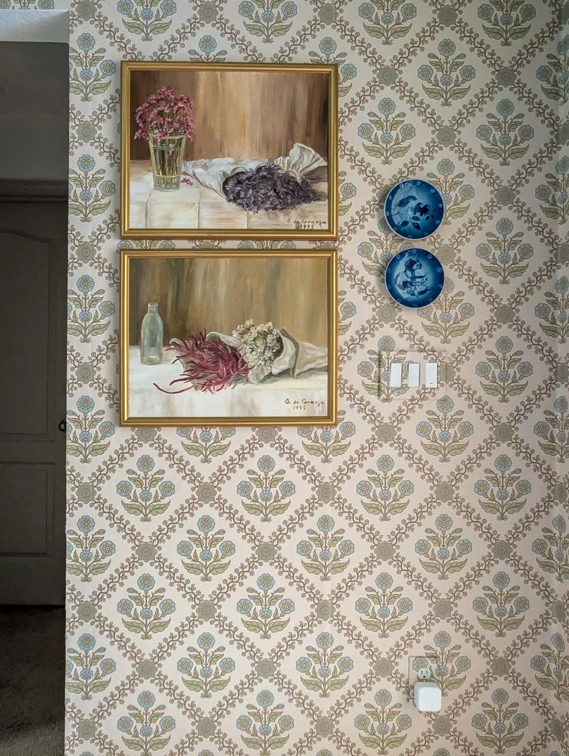 A close-up shot of a vintage-inspired gallery wall features two gold-framed oil paintings of floral still lifes styled alongside blue and white porcelain plates against the intricate pattern of the "Floral Legacy" wallpaper.