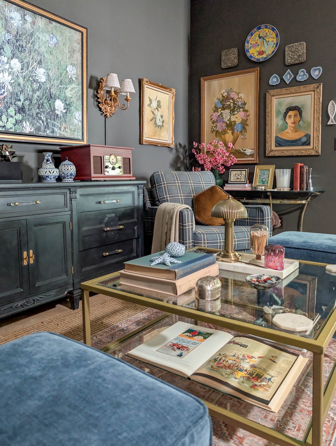 A moody, maximalist living room featuring charcoal walls adorned with gold-framed vintage art and a black sideboard, centered by a blue plaid armchair and a glass coffee table.