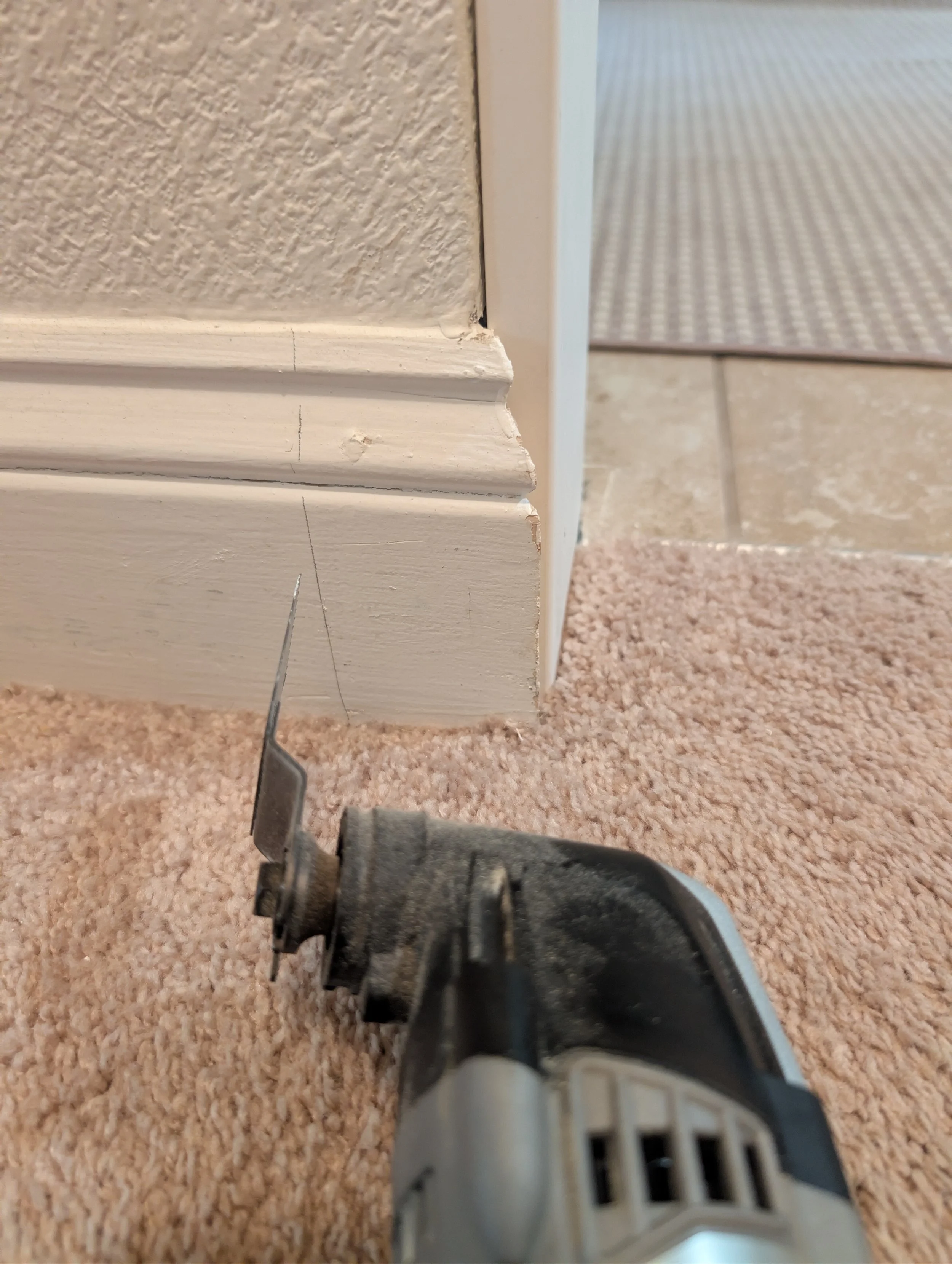 An oscillating multi-tool is positioned on a carpeted floor to cut a vertical line into a white baseboard.