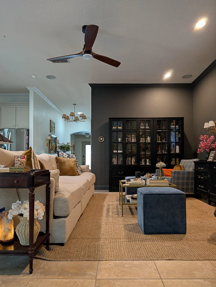 A wide-angle view of a living room with a cream sofa and blue velvet ottoman, featuring a dark charcoal accent wall with a black glass-front cabinet and a wooden ceiling fan overhead.