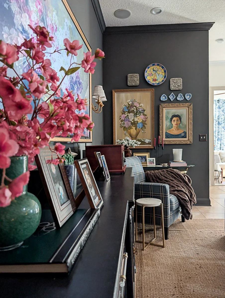 A moody, eclectic living room features dark charcoal walls decorated with a gallery of vintage-style artwork, gold sconces, and blue plaid armchairs arranged around a natural jute rug.