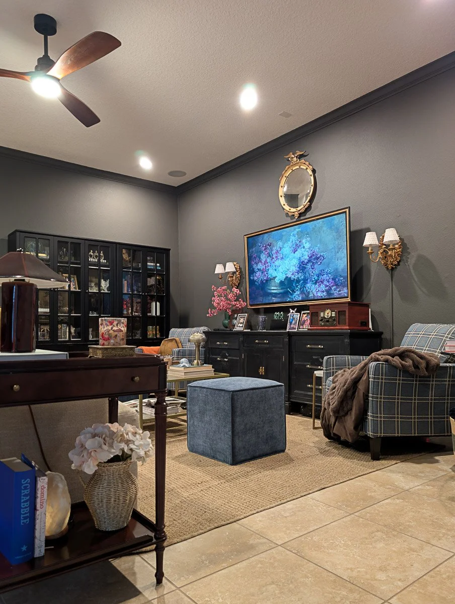 A cozy sitting area with dark grey walls, featuring a large floral digital art piece framed on the wall, two blue plaid armchairs, and golden wall sconces.