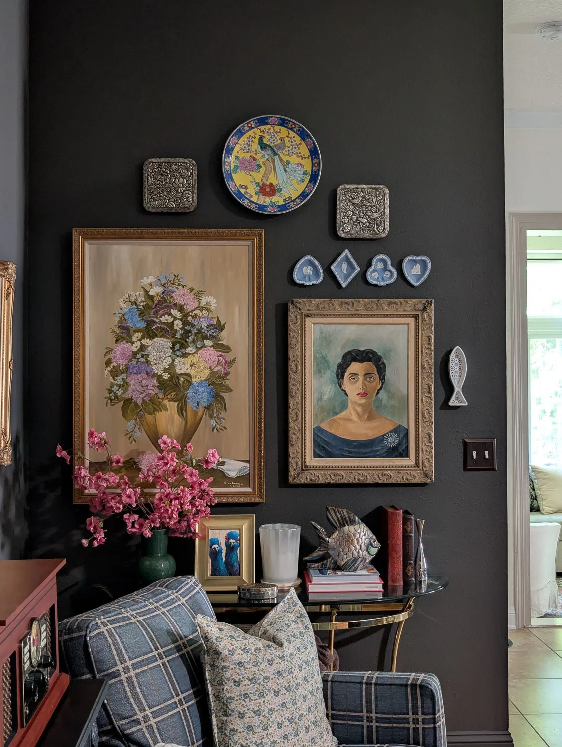 Close-up of a moody traditional gallery wall featuring an oil painting of a woman in a blue dress, a floral oil painting, and vibrant pink faux cherry blossoms in a dark green glass vase on a sleek console table.