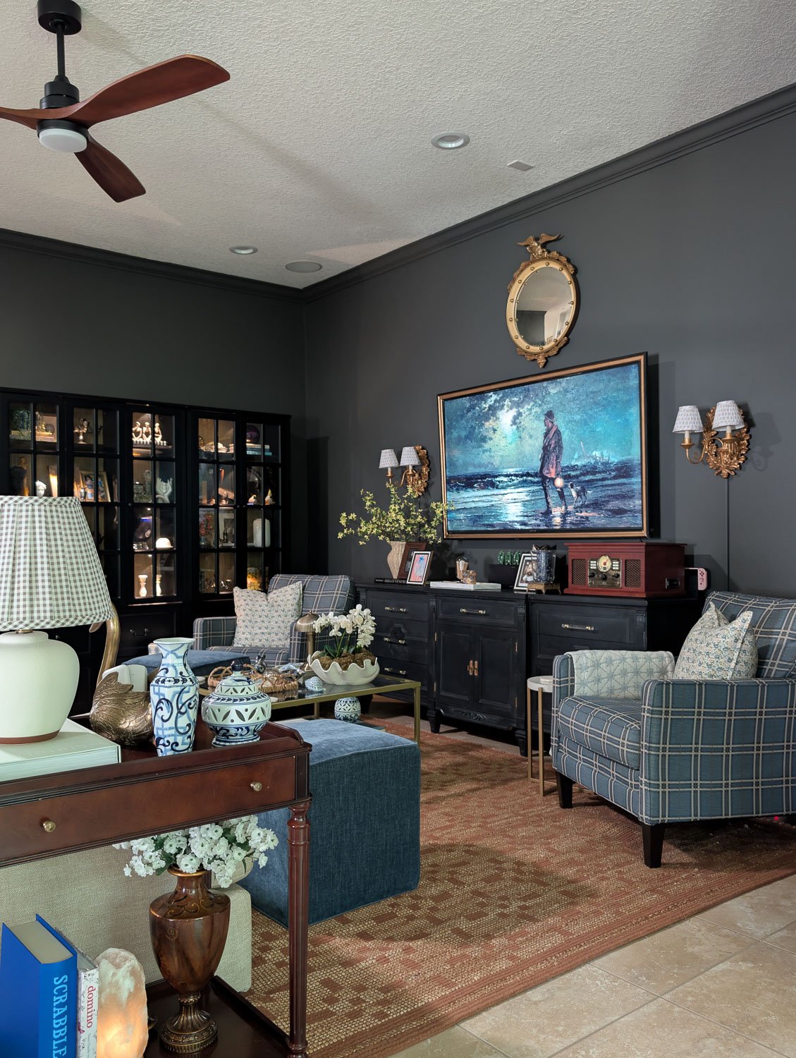 An angled view of a styled moody traditional living room with dark gray walls, blue plaid accent chairs, a terracotta patterned area rug, a brass-framed glass coffee table, and an antique gold convex mirror hanging above a frame TV.
