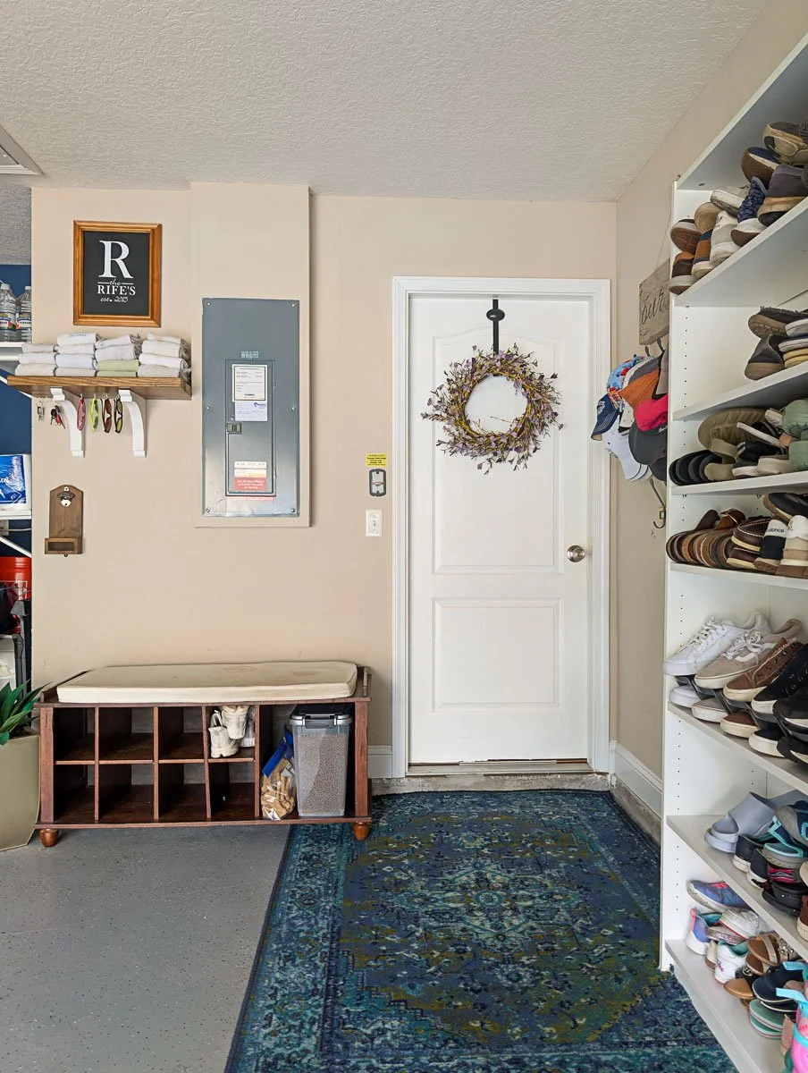 A straight-on view of the garage entry features a white door with a purple wreath, flanked by a shoe bench on the left and a large white shoe shelf on the right.