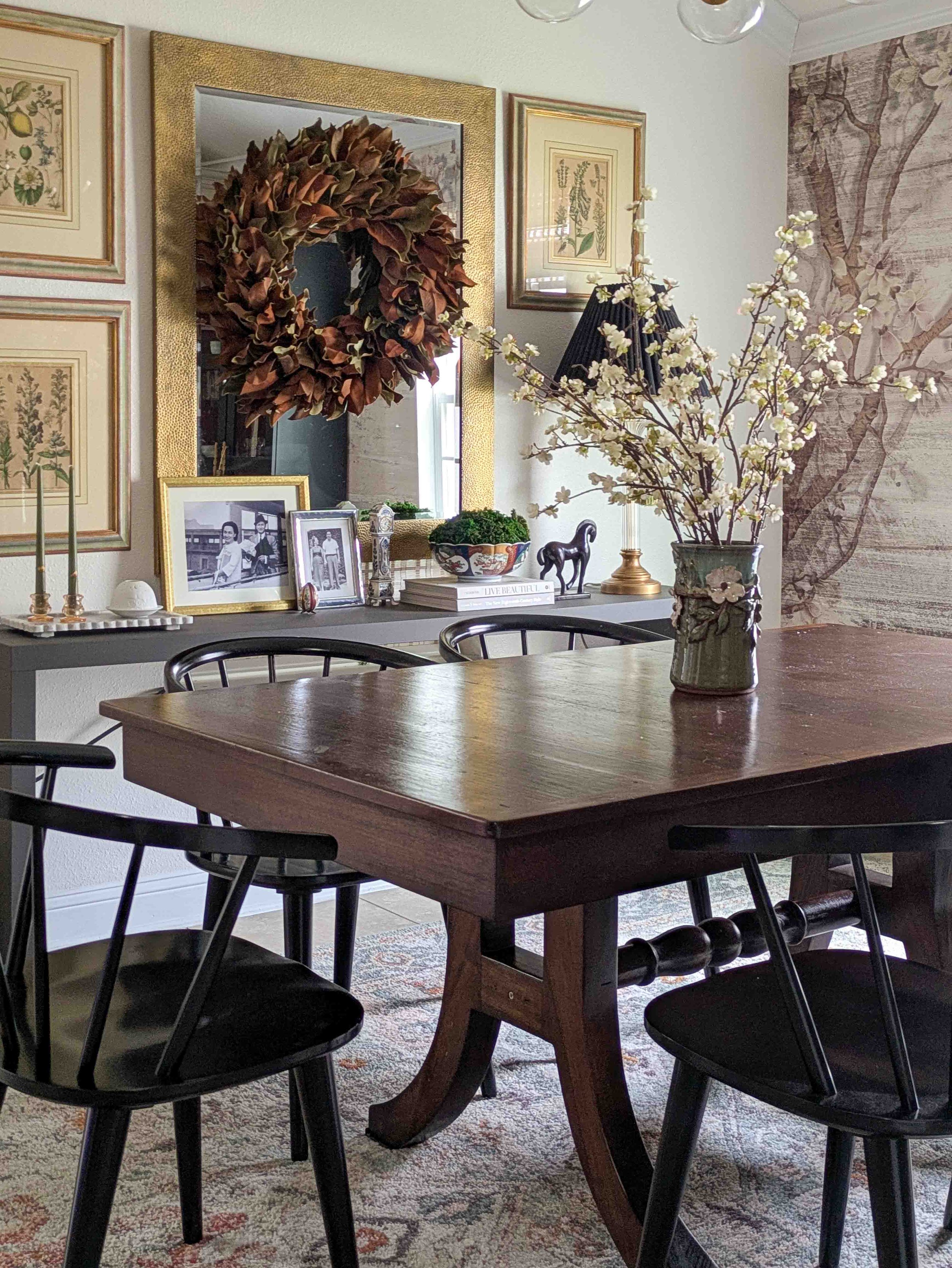 A dark wood dining table with a floral vase in the foreground, backed by a console table styled with a large gold-framed mirror, a magnolia wreath, and intentional book stacks.