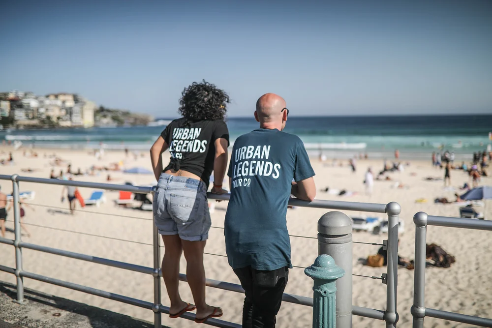 Two of our crew in Urban Legends shirts leaning on the Bondi Beach railing, looking out over the sand and surf