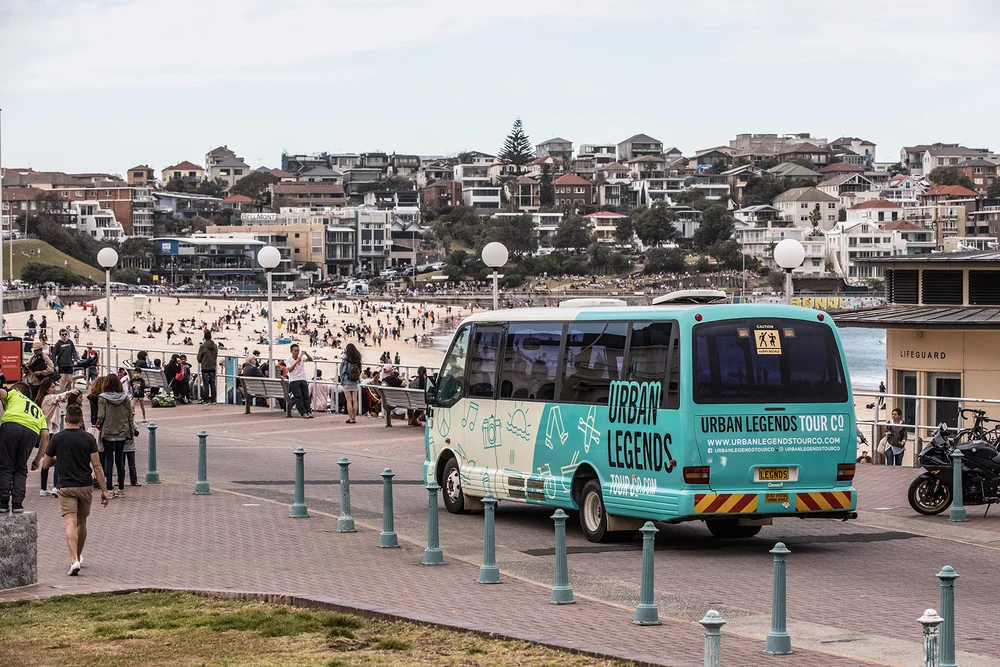 An Urban Legends minibus on the Bondi Beach promenade with the beach and the Bondi hillside behind