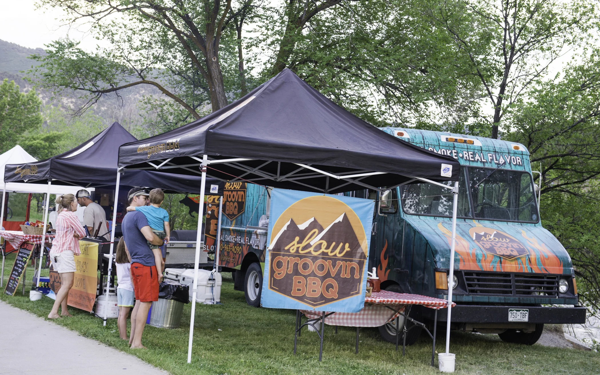 A food truck with a sign that reads "slow groovin BBQ" parked with tents set up for a barbecue event, people waiting in line and interacting near the food truck outdoors.