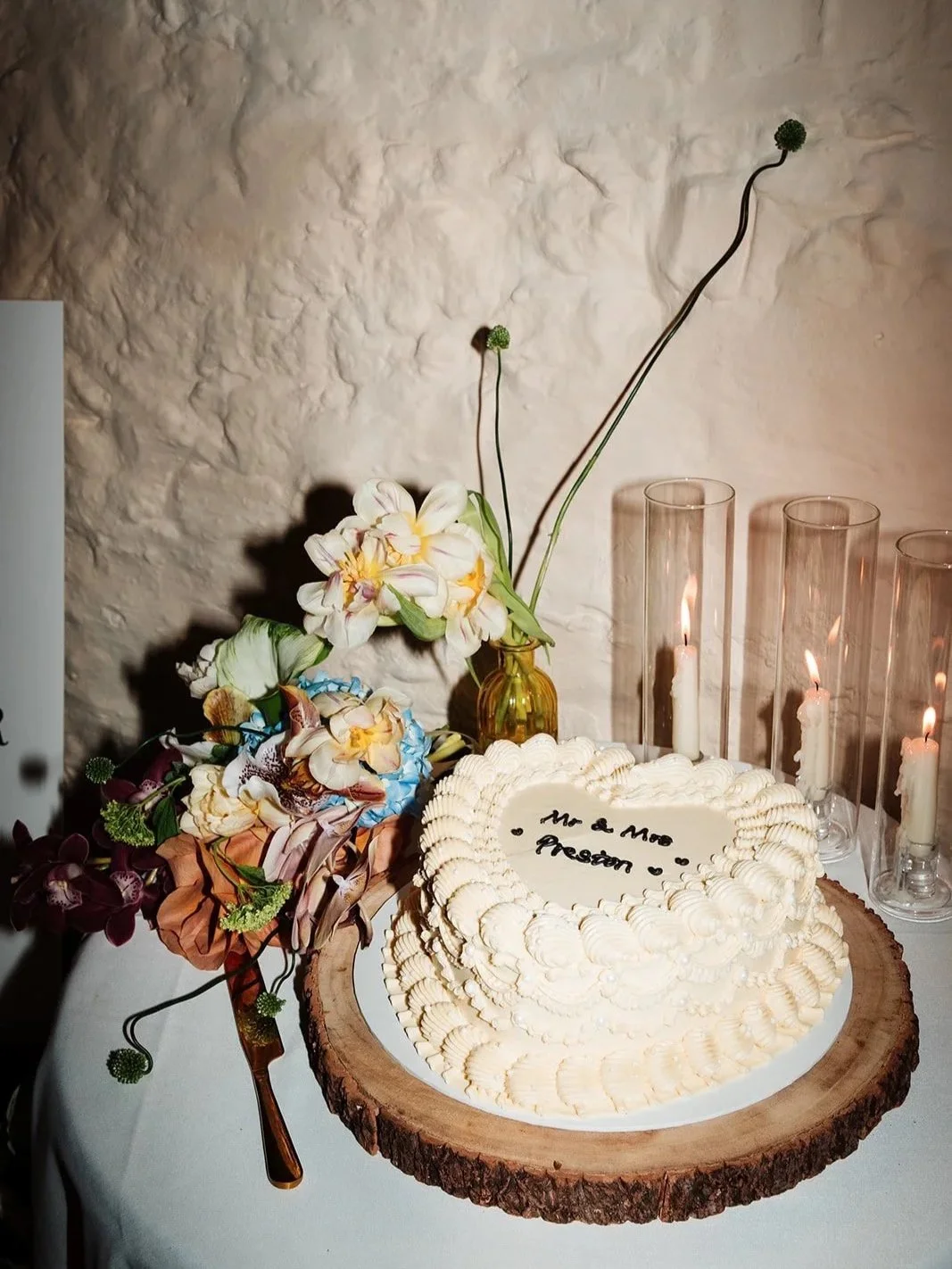 A wedding cake with white icing and floral decorations, topped with handwritten message in Italian, on a wooden slab with flowers and candles nearby.