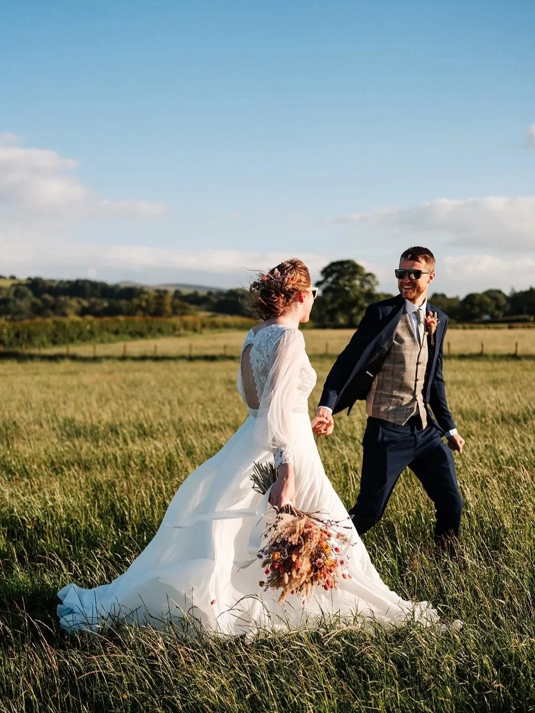 A bride and groom walking hand in hand across a grassy field on a sunny day, the bride in a white wedding dress holding a bouquet, and the groom in a suit with sunglasses, smiling at each other. The wedding is at Low Hall in the Lakes.