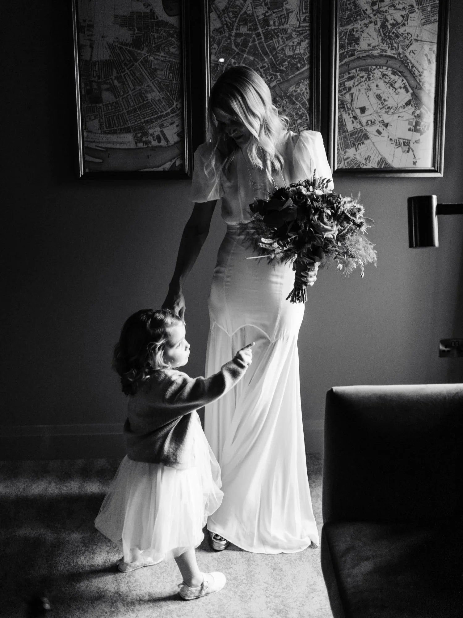 A woman in a long wedding dress holding a bouquet of flowers, standing in front of a young girl, both inside a room with framed maps on the wall. The woman bends slightly downward, and the girl, dressed in a tulle skirt and sweater, points her finger up towards the woman.