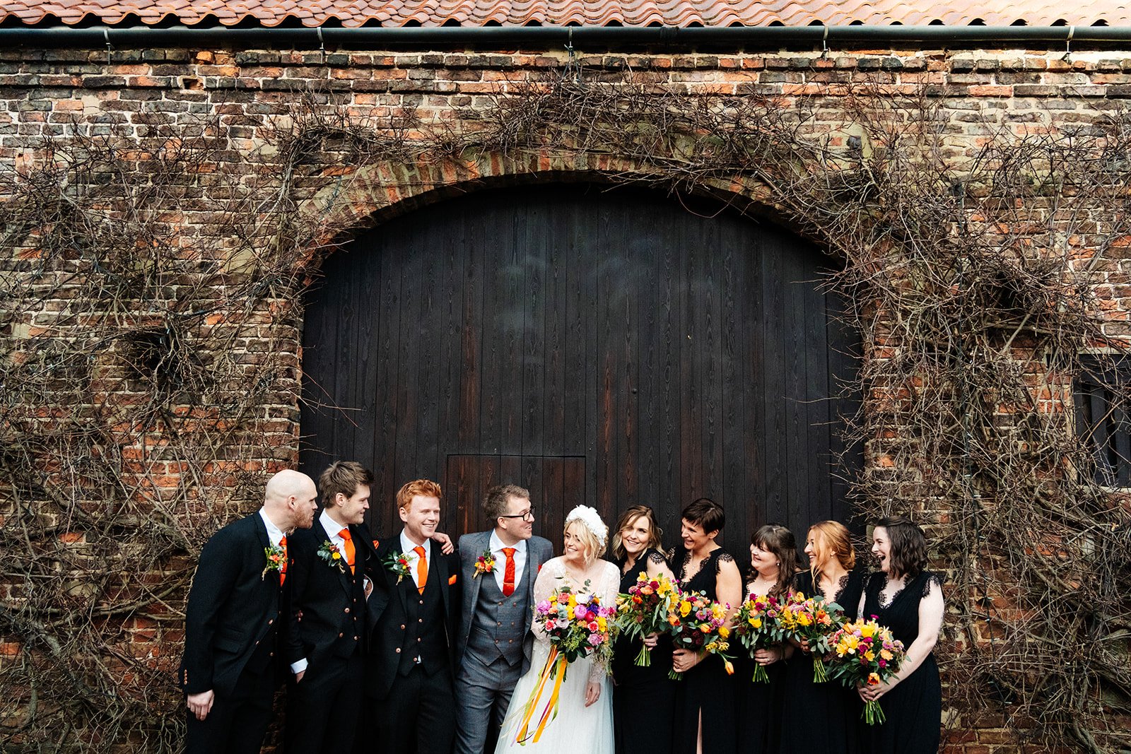 group shot of bride, groom, bridesmaids and groomsmen. The woman are wearing black dresses and have colourful flowers. Everyone laughing. the normans York wedding