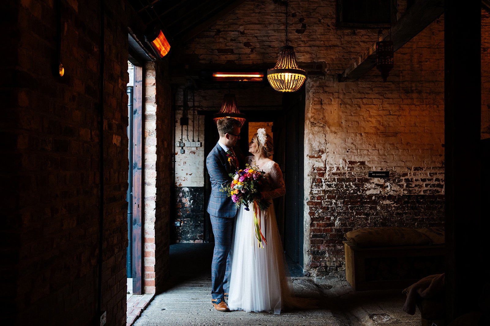 bride and groom stand for a portrait in a side-lit barn room. Colourful flowers. the normans York wedding