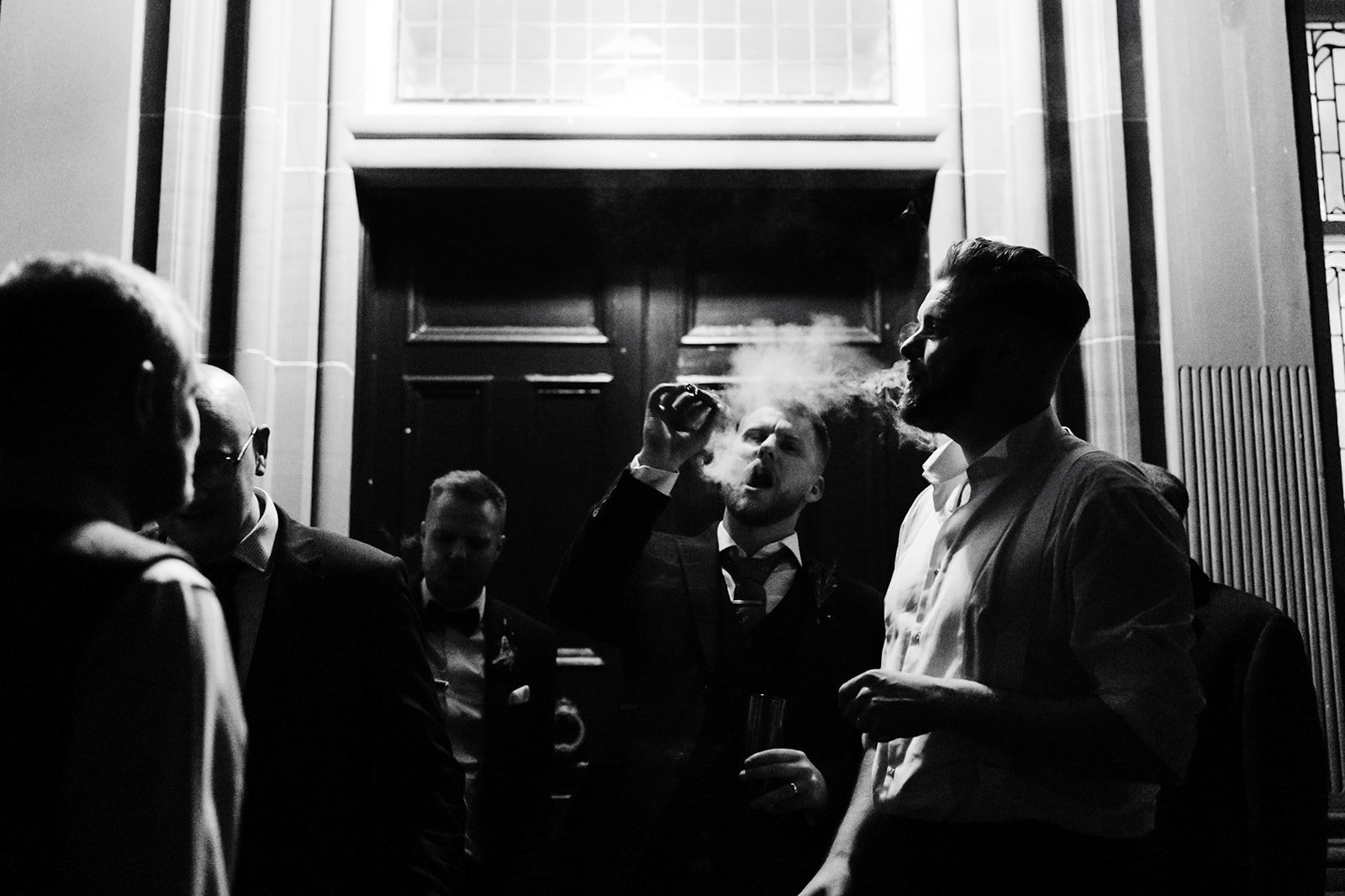 groom and his friends smoke cigars, black and white, in the porch of a mansion house. a smokey atmospheric shot of the bride and groom cheering and dancing. wedding at toftcombs in Scotland