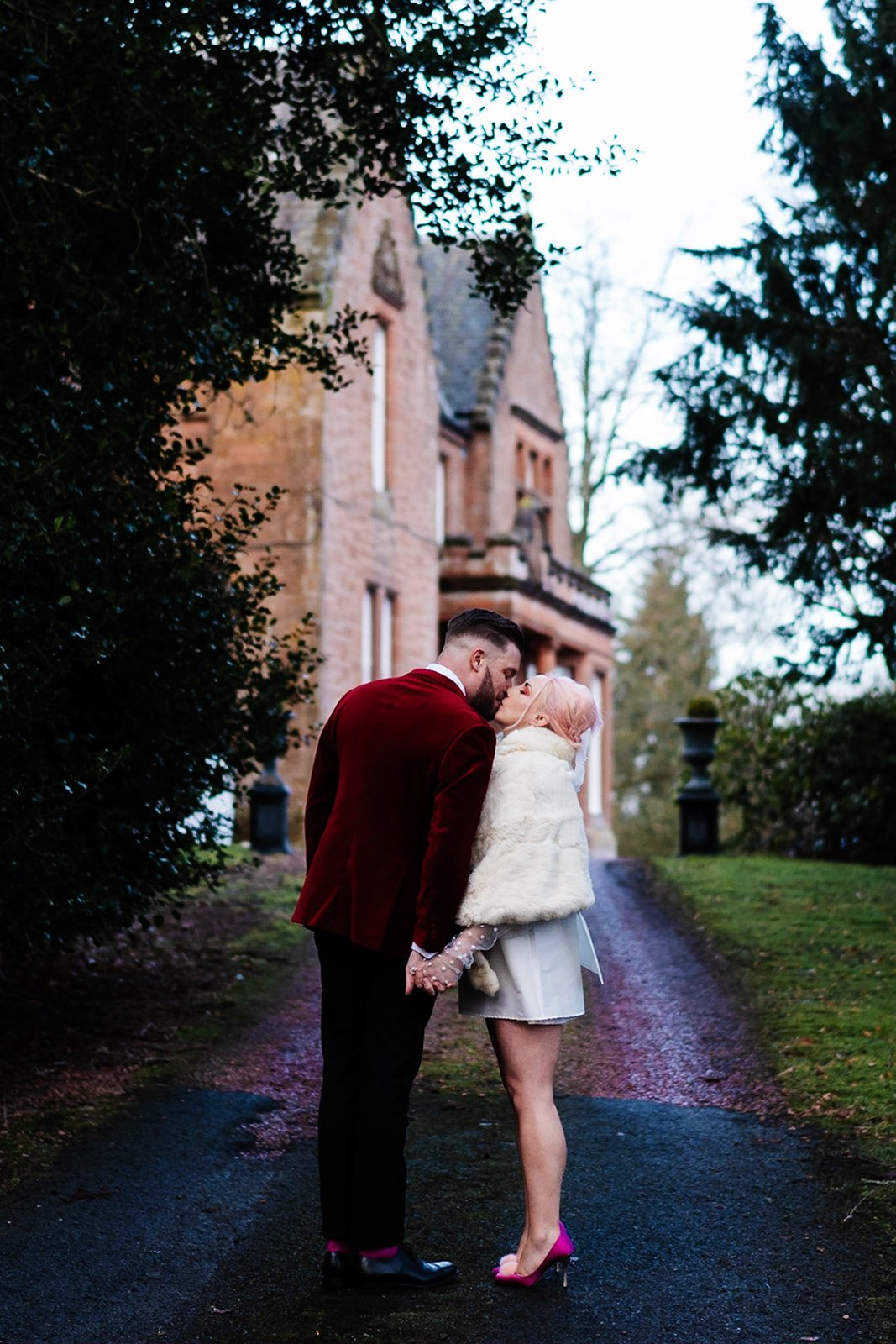 bride and groom kiss with toftcombs house in the background. red velvet suit jacket