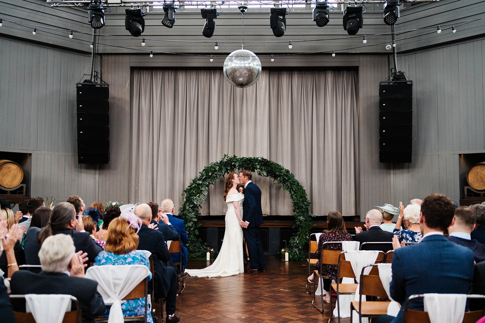 A bride and groom kiss during the ceremony under a botanical arch and a mirror ball at Wylam Brewery in Newcastle. North-east wedding photography