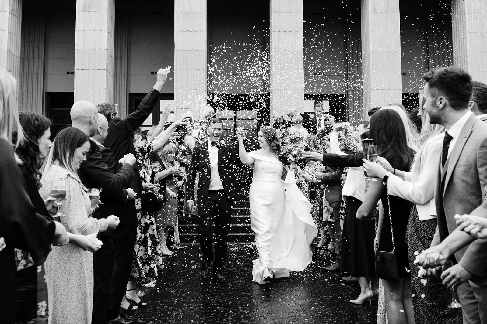 Black-and-white image of a bride and groom cheering as they walk through confetti outside Wylam Brewery in Newcastle. North-east wedding photography