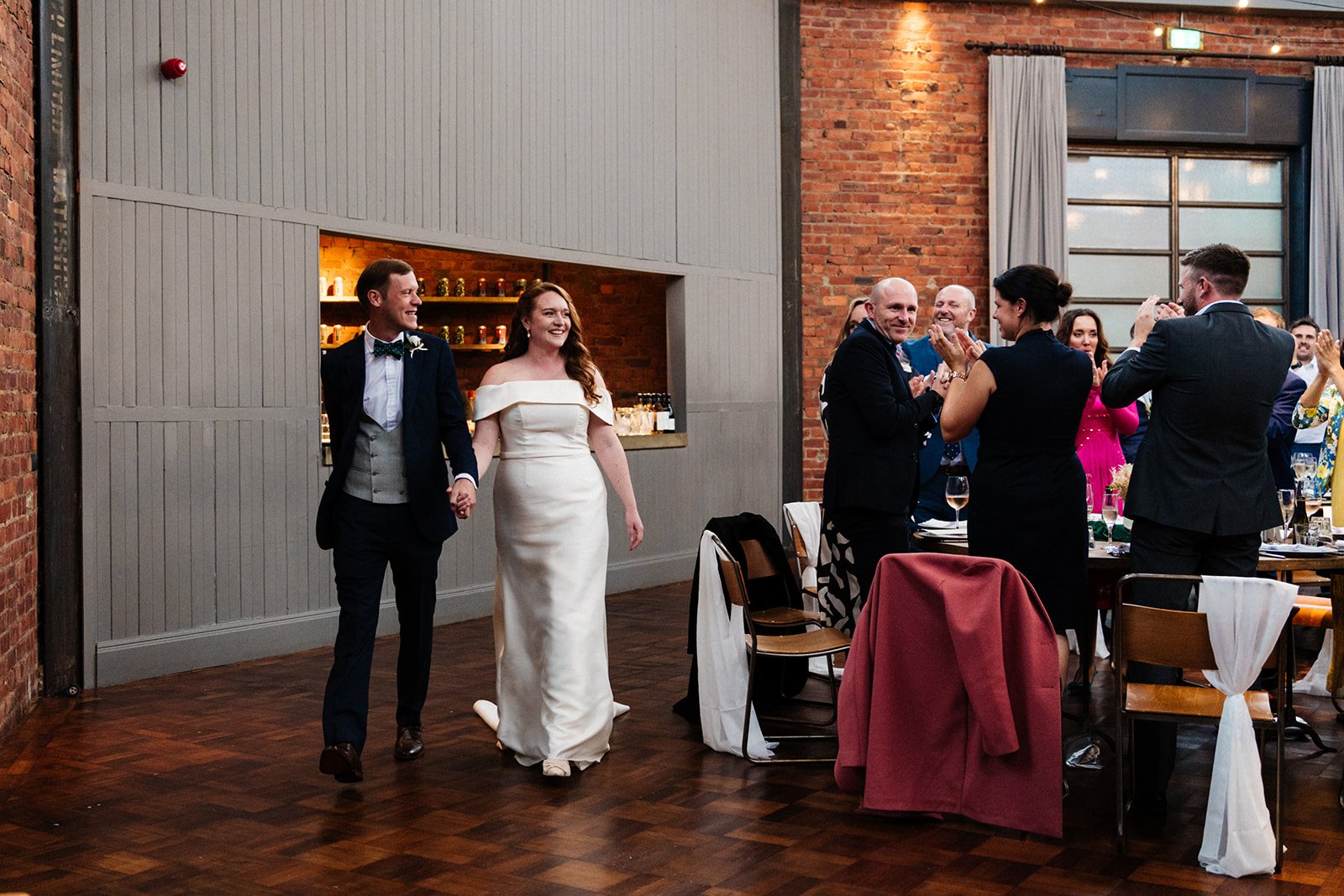A bride and groom smile their guests as they walk into the mail room at Wylan Brewery in Newcastle ahead of their wedding speeches