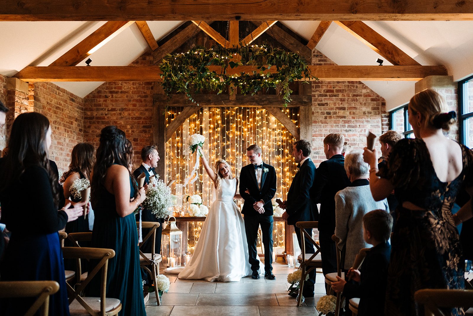 The bride and groom are at the end of the wedding aisle and the bride is holding her bouquet up and cheering. Wedding at Thirsk Lodge Barns