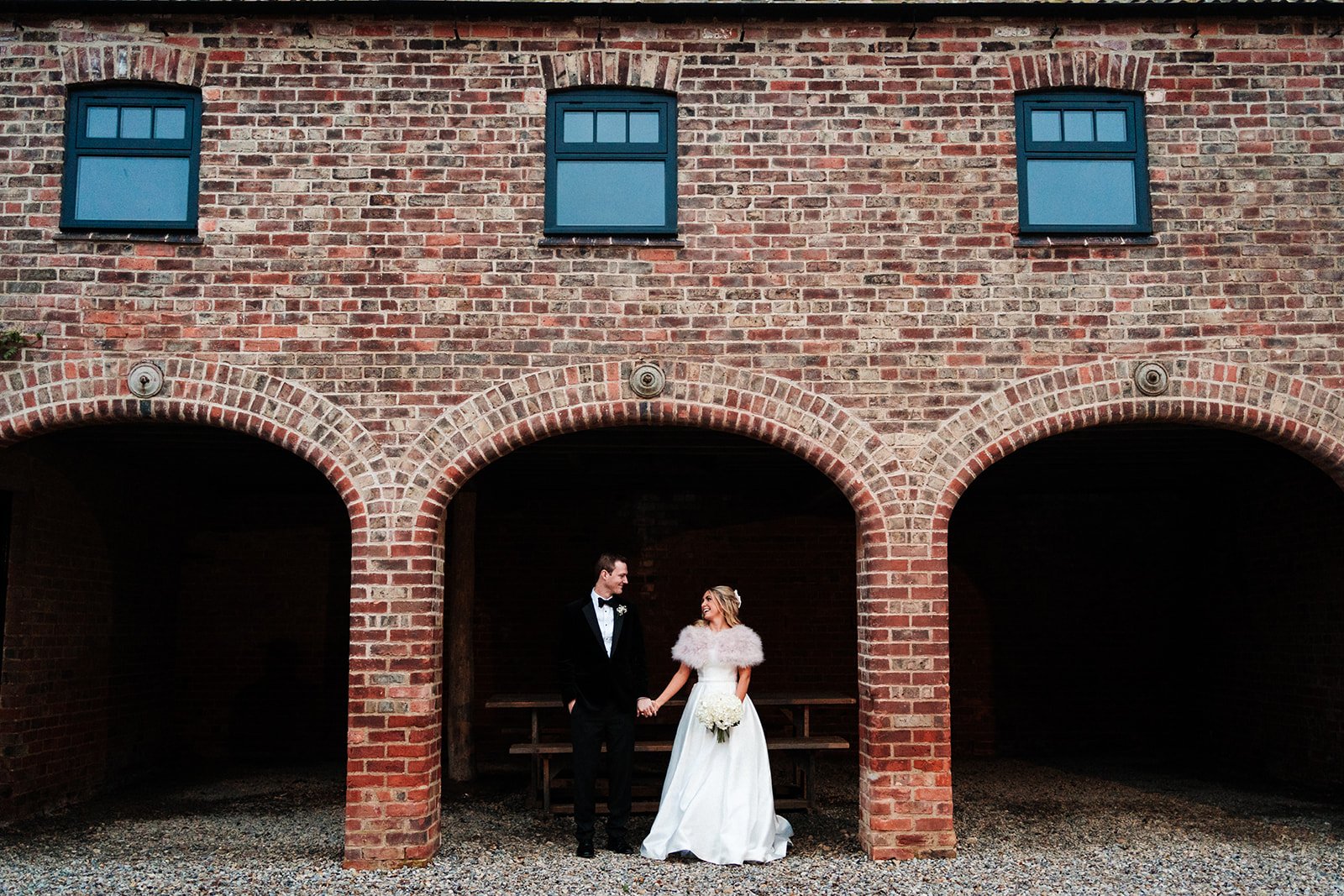 A bride and groom stand hand-in-hand under the archway of a large barn building