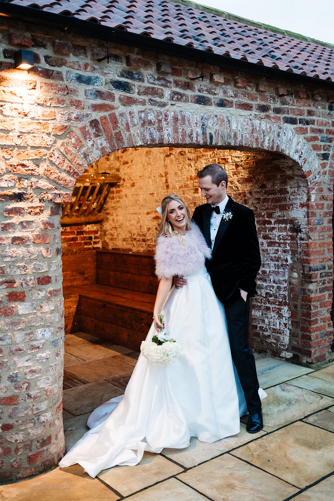 Bride and groom stand close next to a barn building and are laughing together