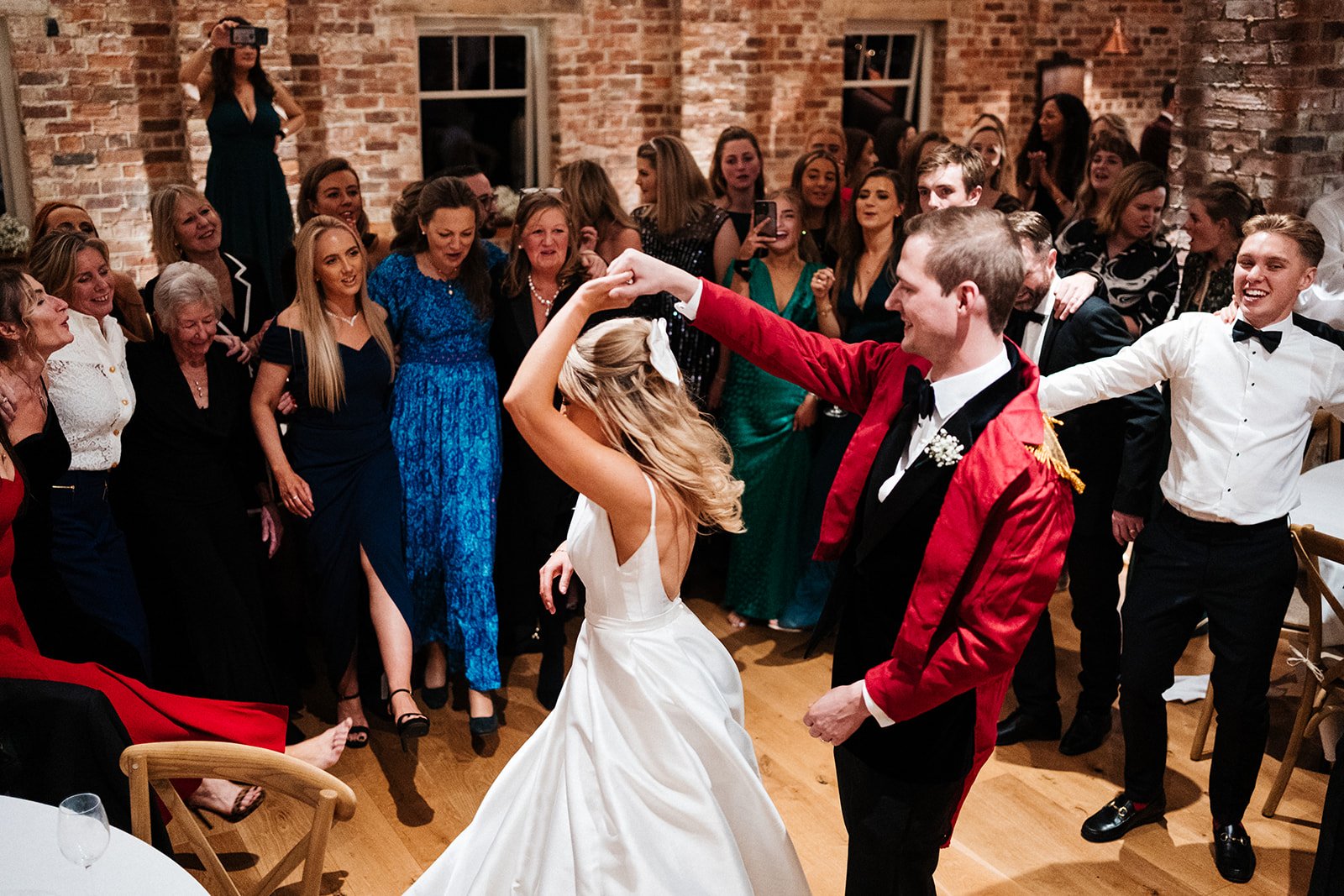 The groom at 12 the bride round surrounded by guests in a barn reception room. Wedding at Thirsk Lodge Barns