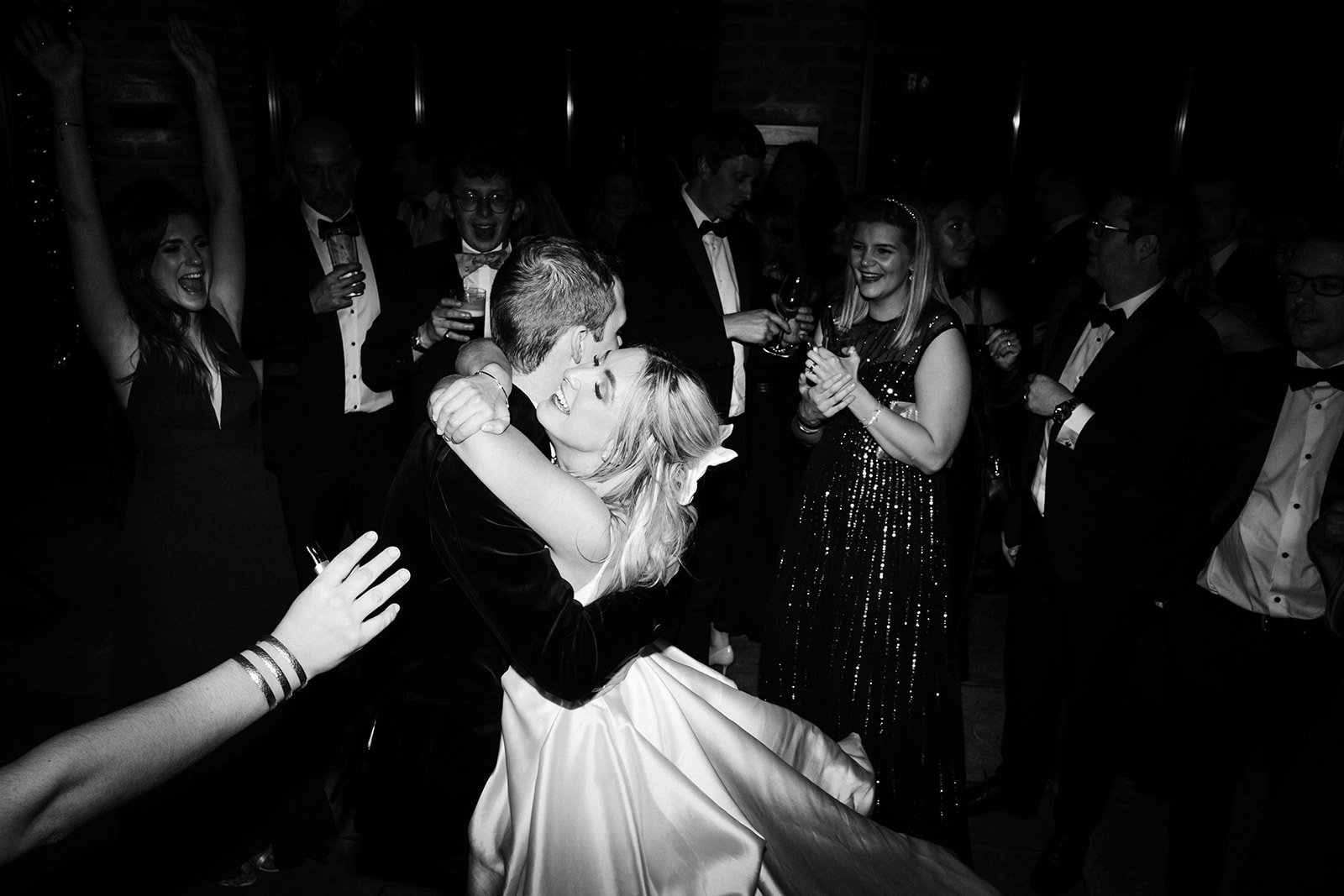Black and white picture of the bride cuddling the groom while dancing surrounded by wedding guests