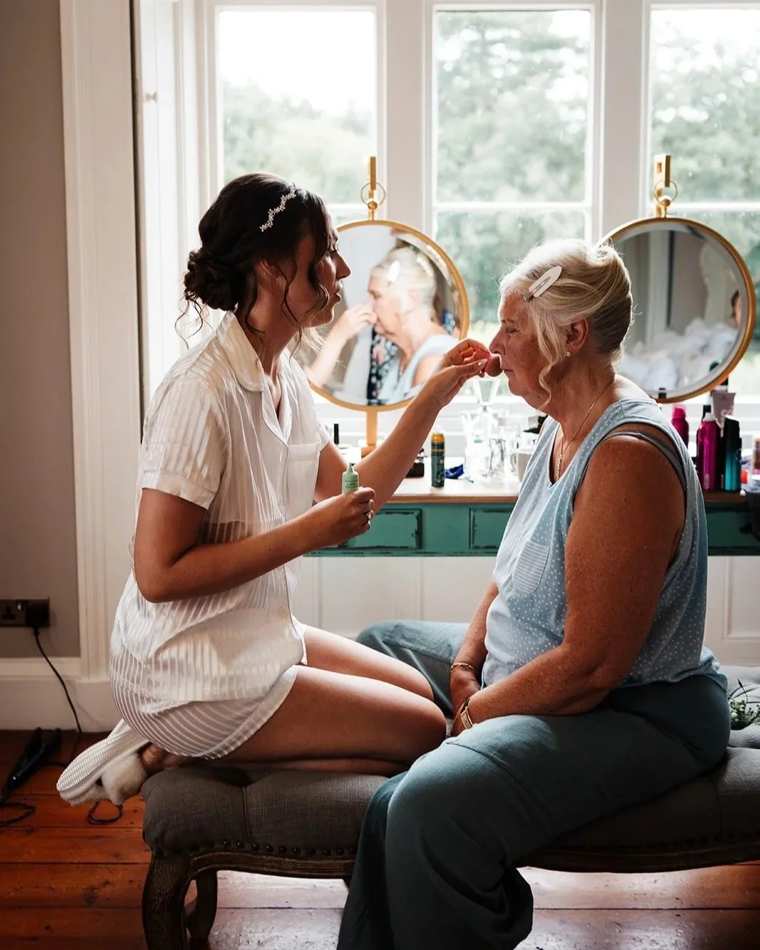A young woman sitting on a cushioned bench in front of an older woman, applying makeup near her nose, in a well-lit room with large windows. This is a wedding at Thicket Priory near York.