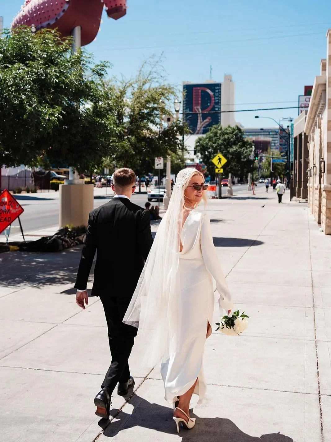 A bride in a white wedding dress and veil carrying a bouquet walks with a groom in a black suit on a city sidewalk on a sunny day. This wedding looks like a Las Vegas elopement.