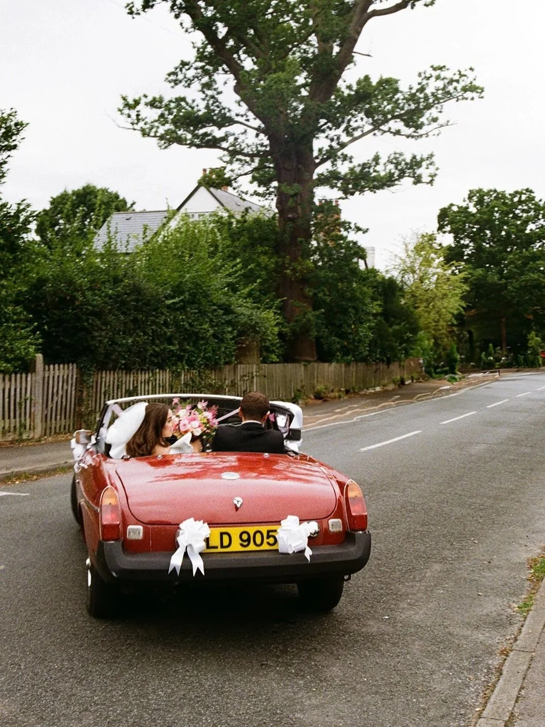 A red vintage convertible car decorated with white ribbon bows, parked on a suburban street. A bride and groom are seated inside, with the bride holding a bouquet of pink flowers, during a wedding celebration. The wedding was shot on 35mm film.