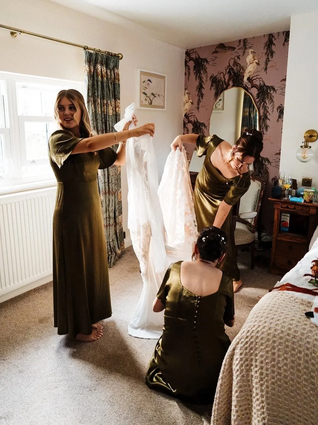 Three women, dressed in matching green satin dresses, preparing a wedding dress in a well-lit bedroom with floral decor. The wedding was shot at Low Hall in the Lakes.