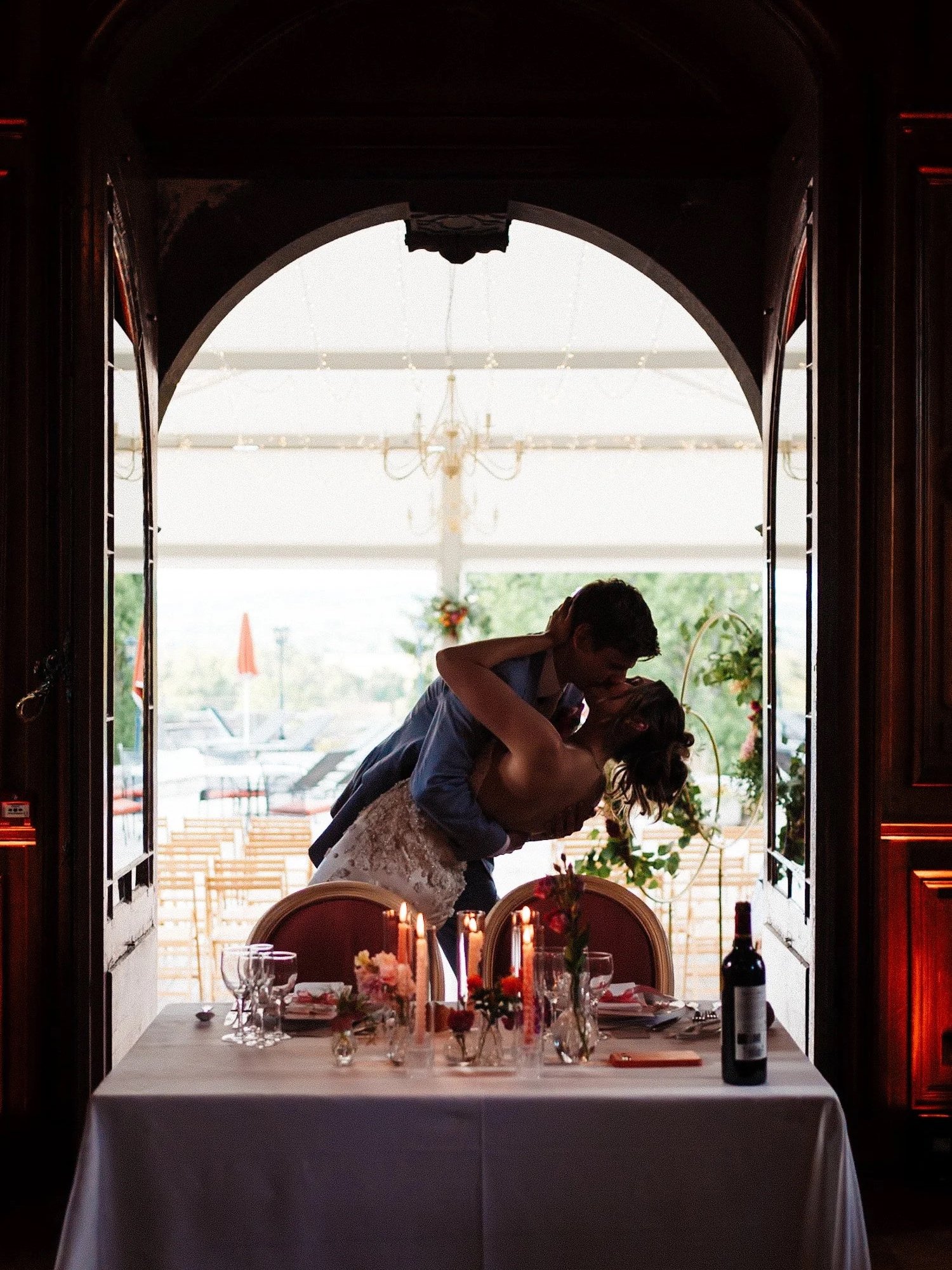 A couple sharing a romantic dance at a wedding reception, standing behind a decorated table with flowers, candles, wine, and glasses, in a room with open doors showing an outdoor patio area. This was a wedding shot at Chateau de Maulmont in France.