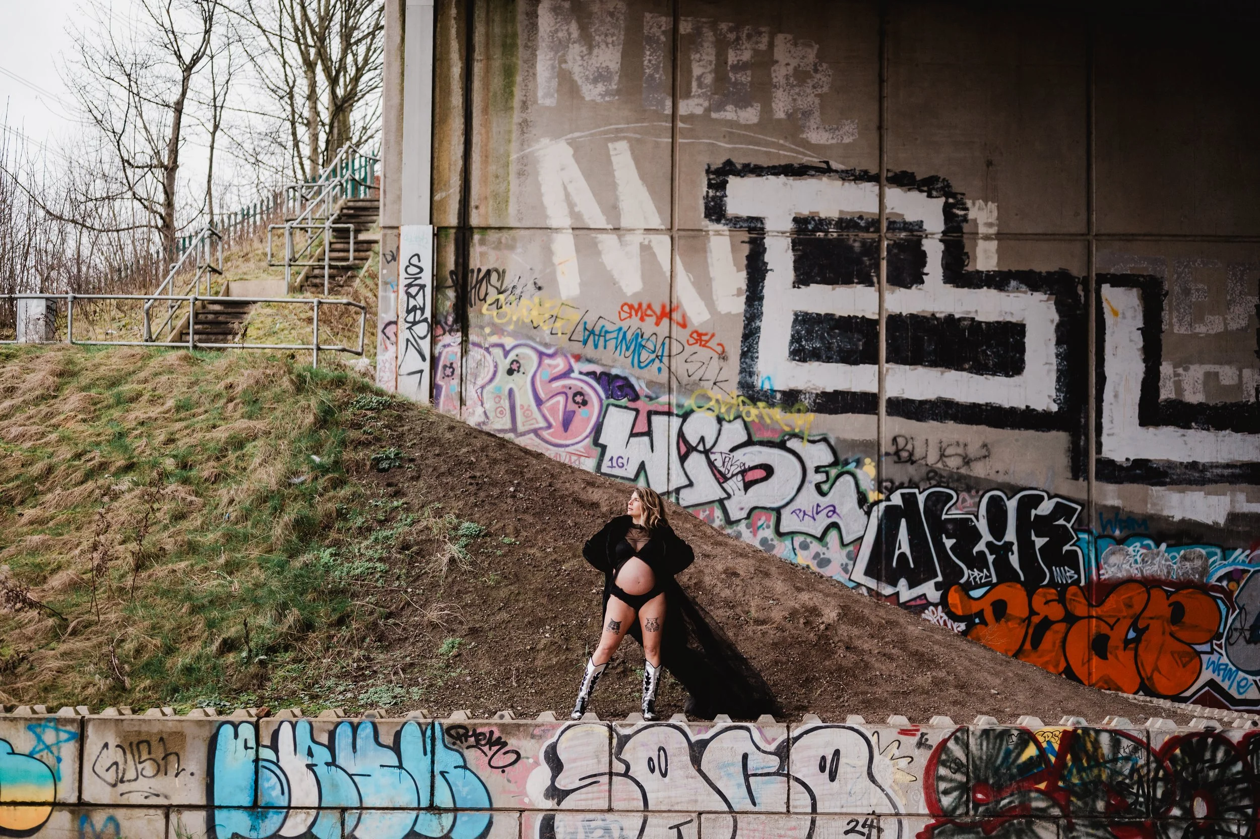 A woman standing on a sidewalk under a graffiti-covered concrete overpass with stairs leading up to a fenced area. She is wearing black lingerie, knee-high boots, and has tattoos on her legs and arms. She is looking up and to her left, with her hands