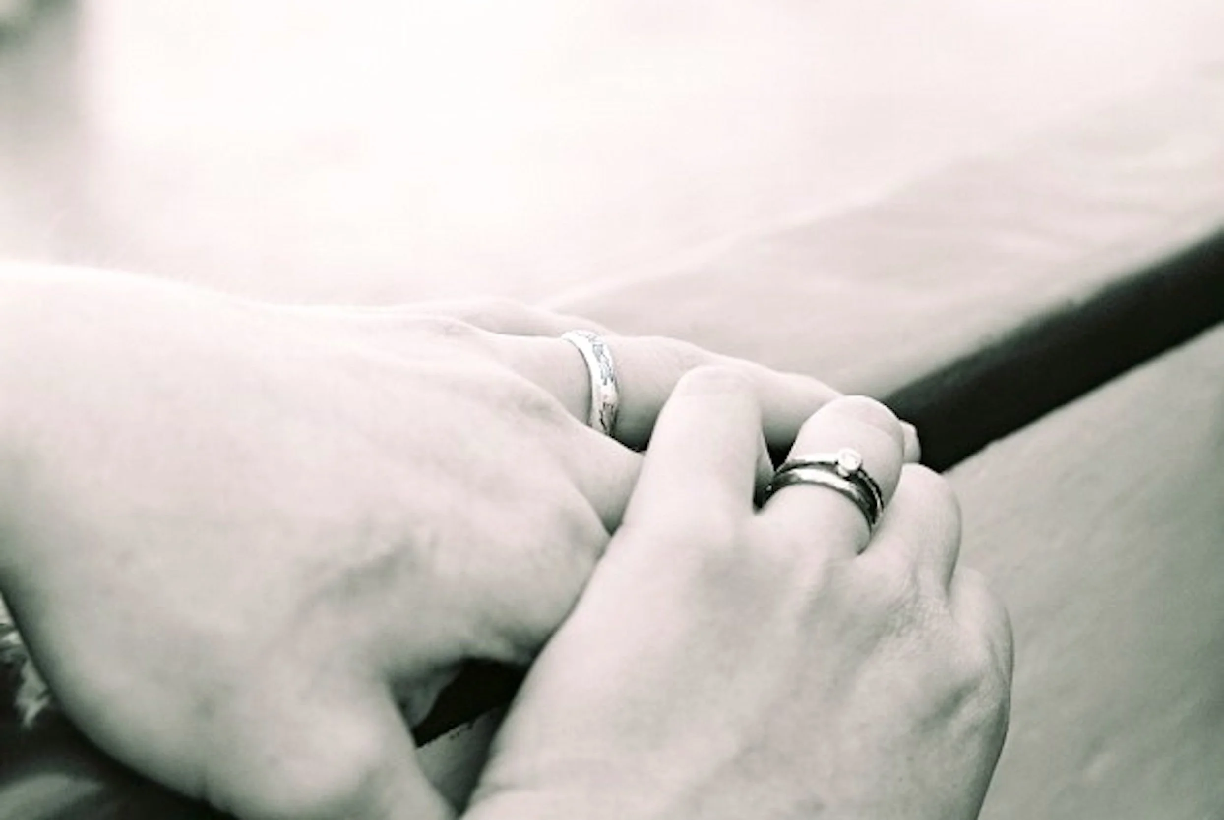 Close-up of two hands holding each other, both wearing rings, with soft focus and black-and-white filter.
