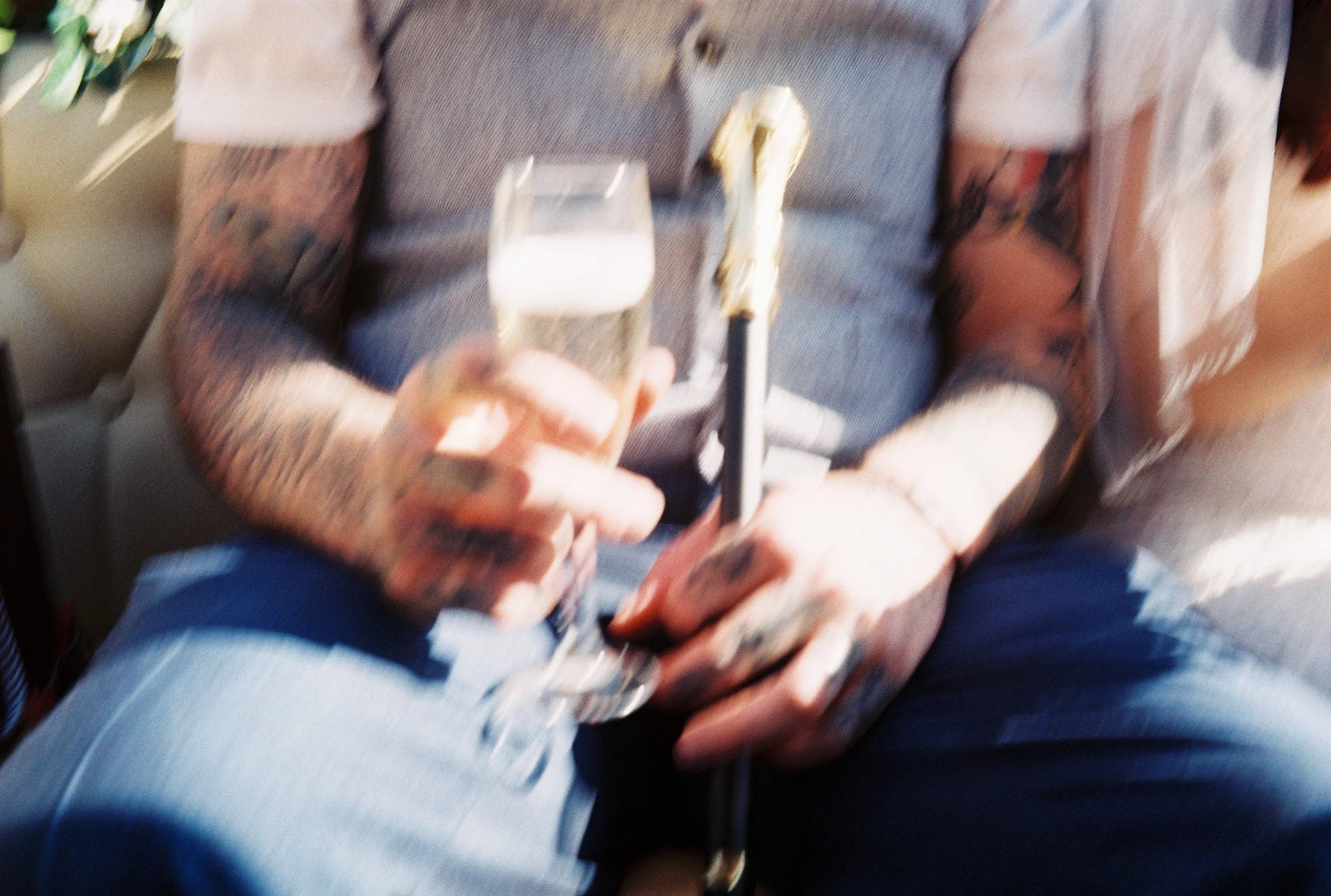 Person with tattoos wearing a gray shirt and blue jeans, sitting and holding a glass of beer and a black smartphone.