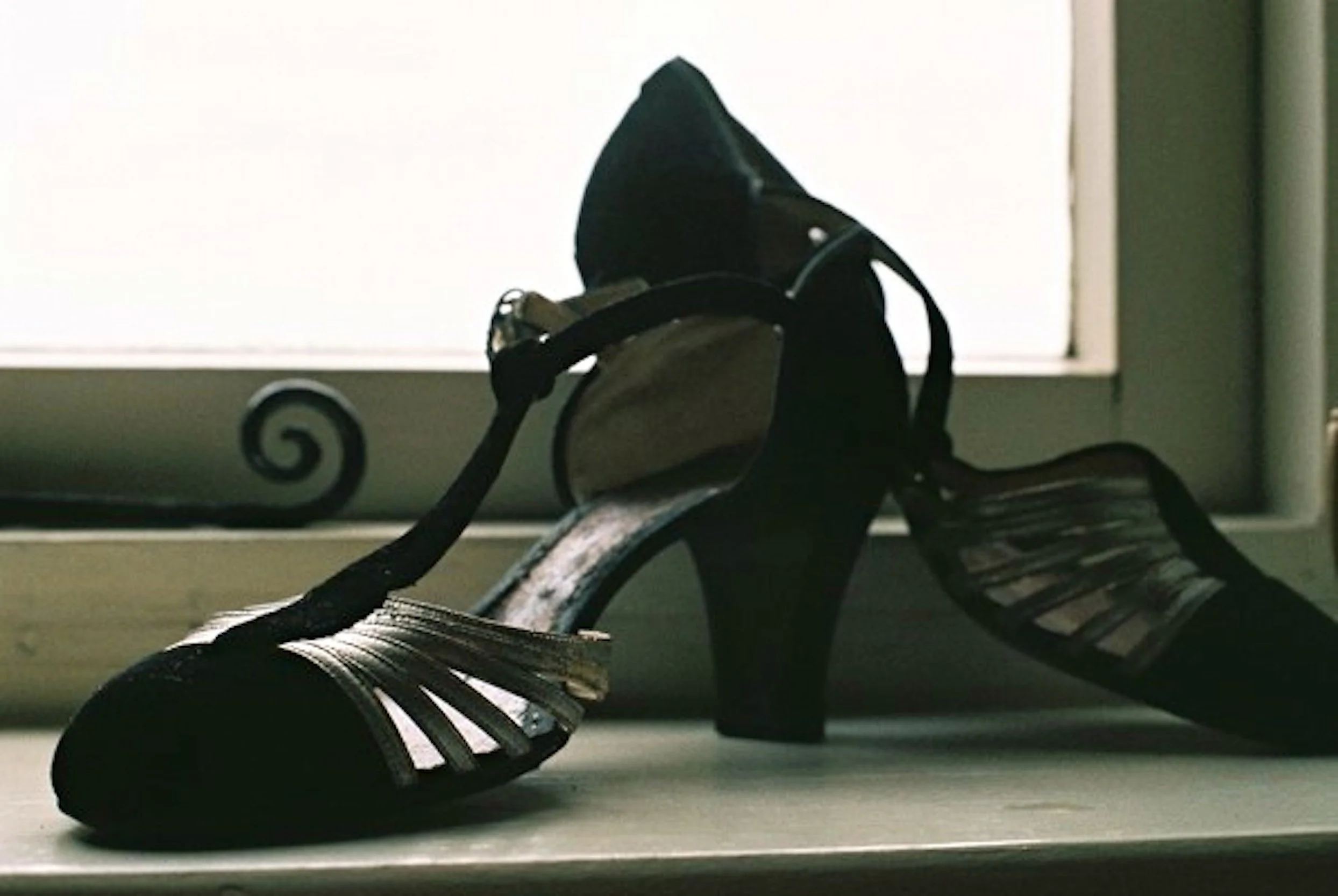 Black high-heeled sandal with wide straps and an ankle strap, placed on a windowsill with frosted window in the background.