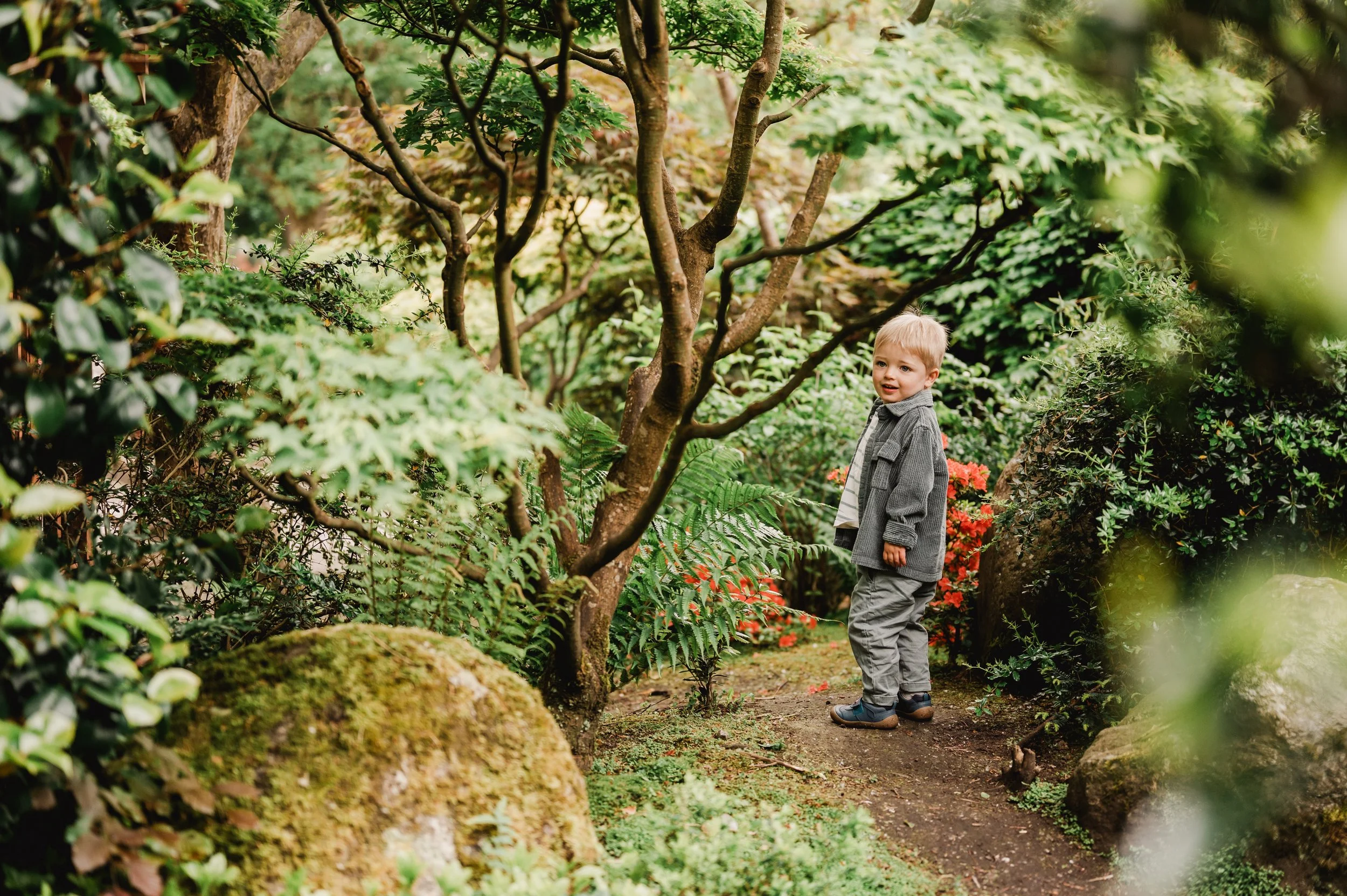 A young boy with blonde hair, wearing a gray jacket and gray pants, walks along a forest path surrounded by lush green plants, trees, and rocks.