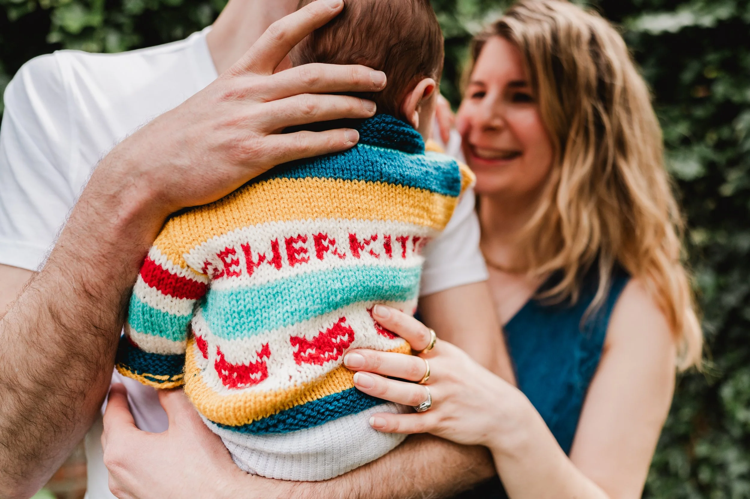 A close-up of a young child wearing a colorful striped sweater with red hearts, being held and cuddled by a woman with long blonde hair, smiling at the child, while someone else gently touches the child's head.
