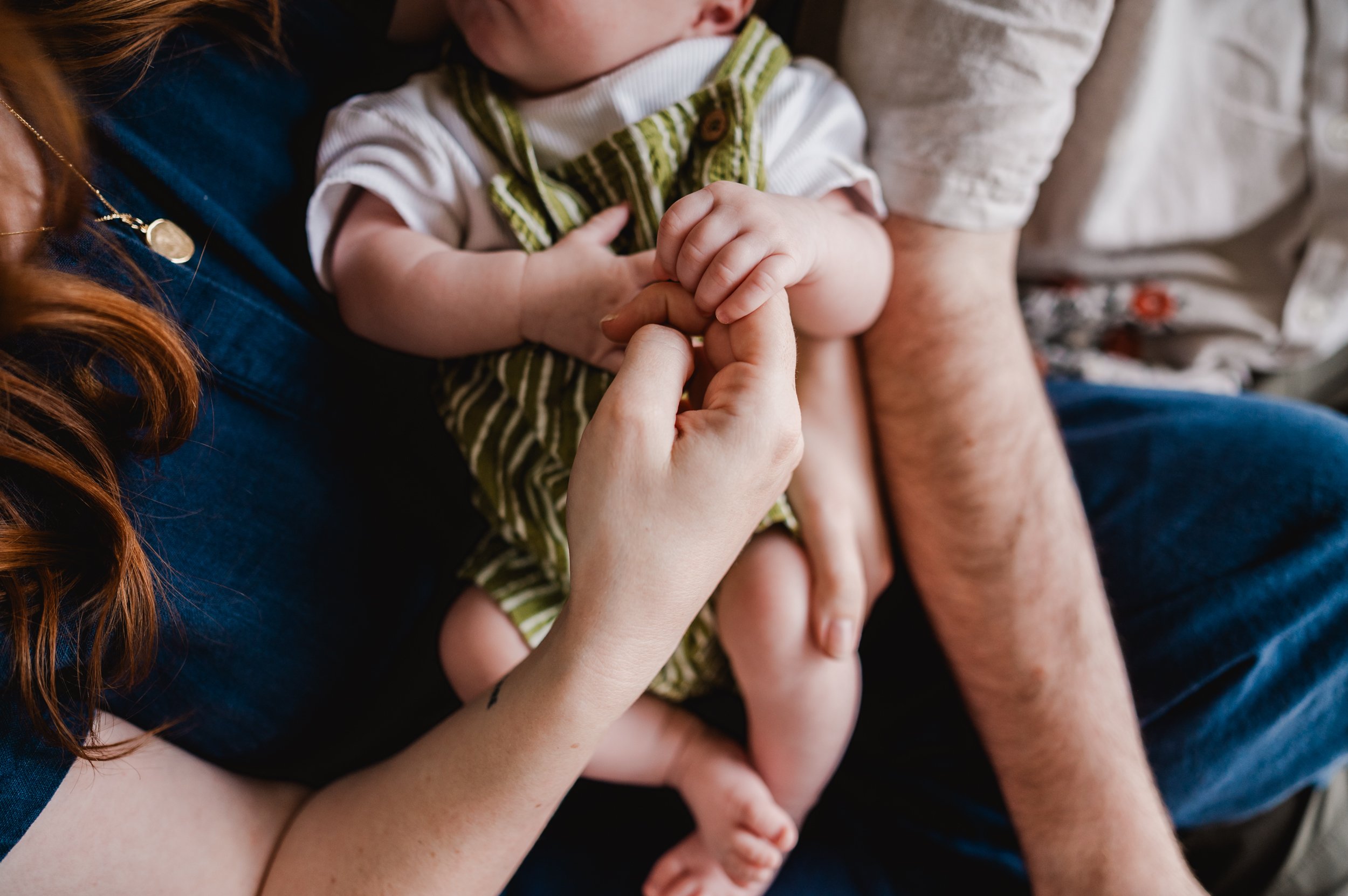 A close-up of a woman with long red hair holding a baby with light skin, wearing a striped green and white outfit. The woman is holding the baby's hand and their faces are close together.