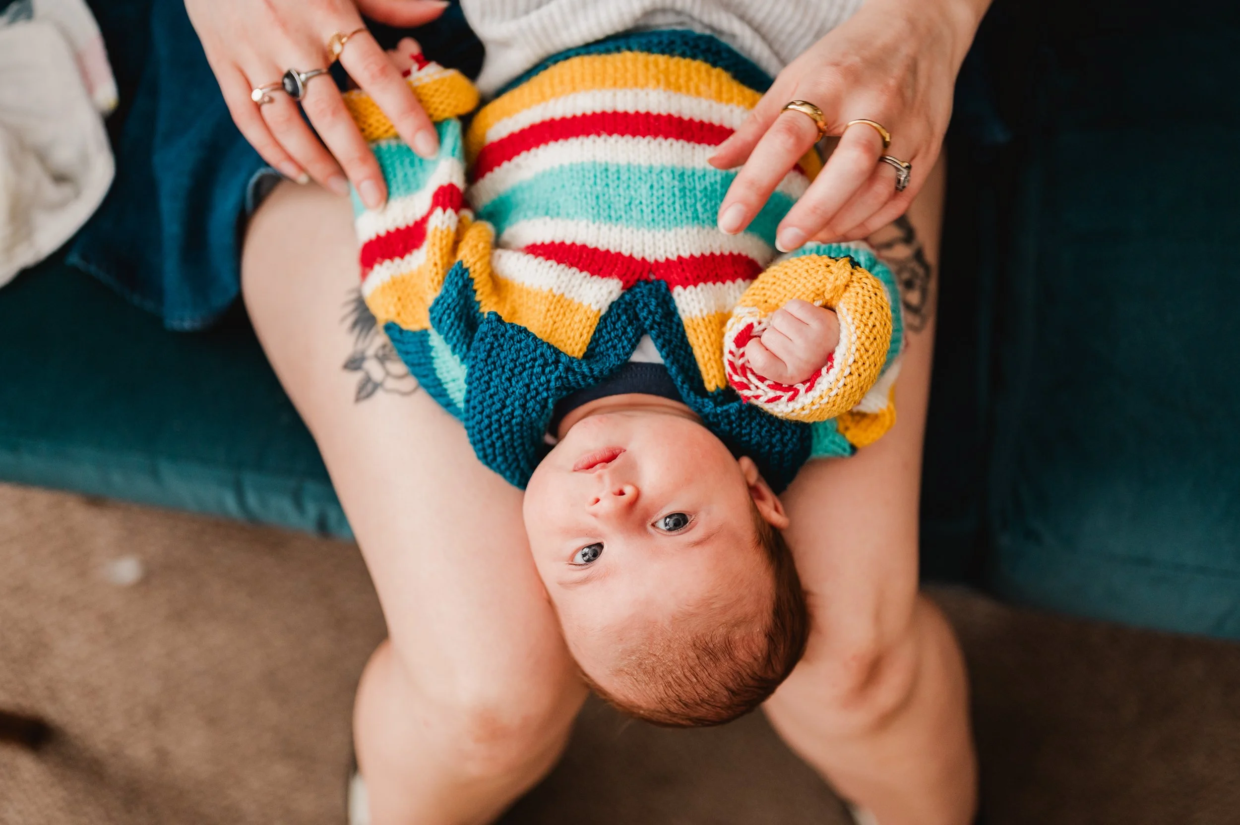 A person holding a baby girl over their chest, with the baby wearing a colorful striped knitted sweater, lying upside down. The baby has blue eyes and light skin, and the person has tattoos, rings, and visible white hair.