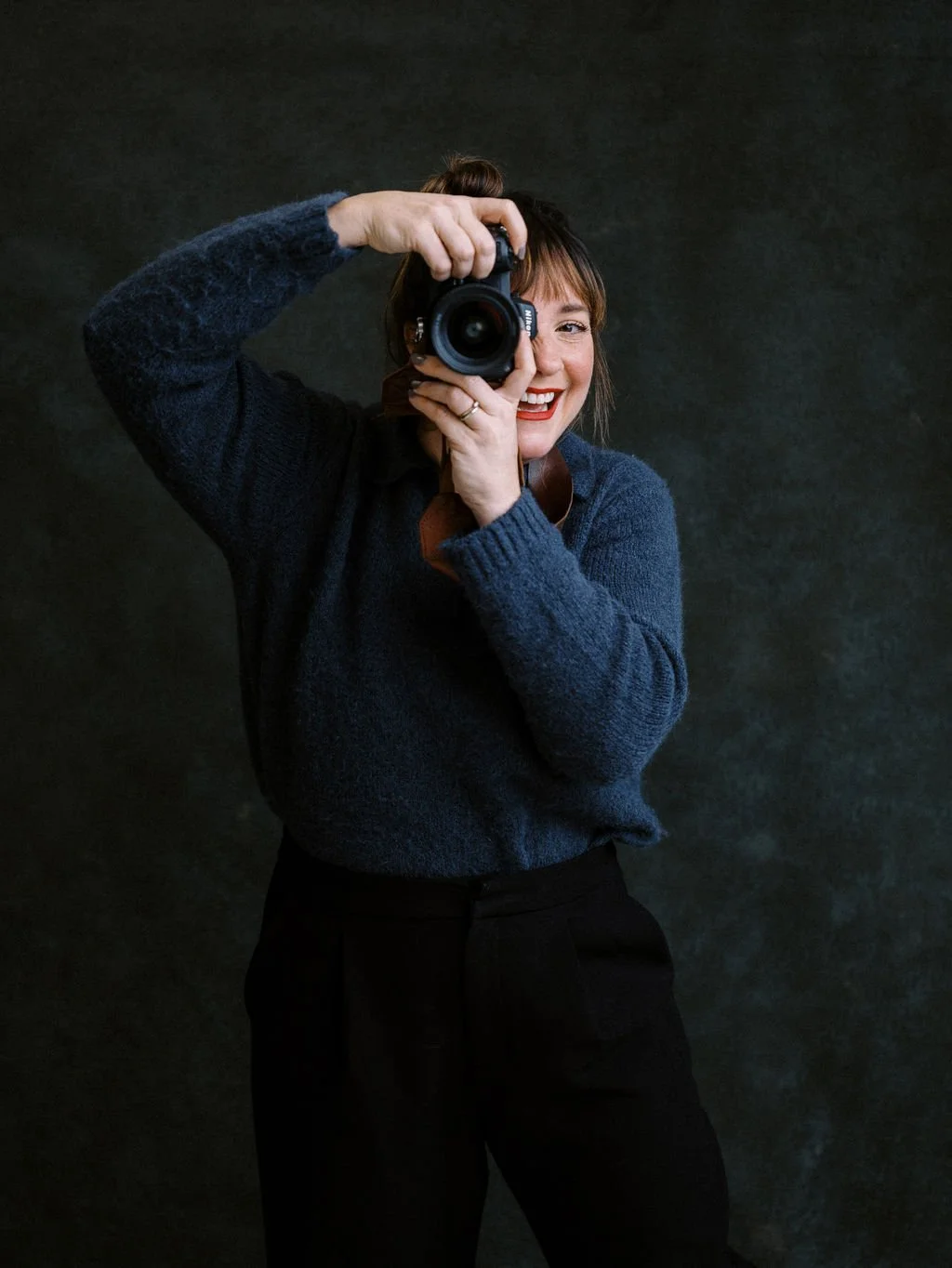 A woman smiling and holding a camera, taking a picture, wearing a dark blue sweater, standing against a dark background.