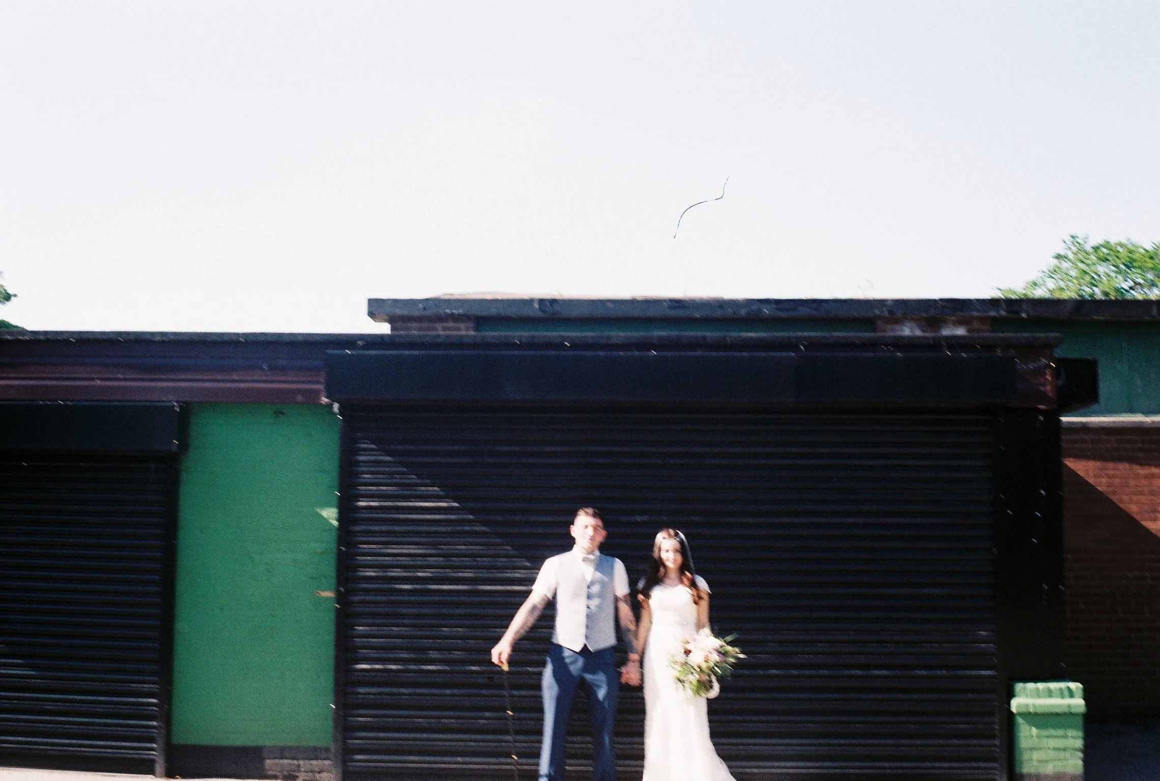 A couple in wedding attire walking hand in hand in front of a black roller shutter garage door, with a green wall and a brick wall on either side, and clear sky above.