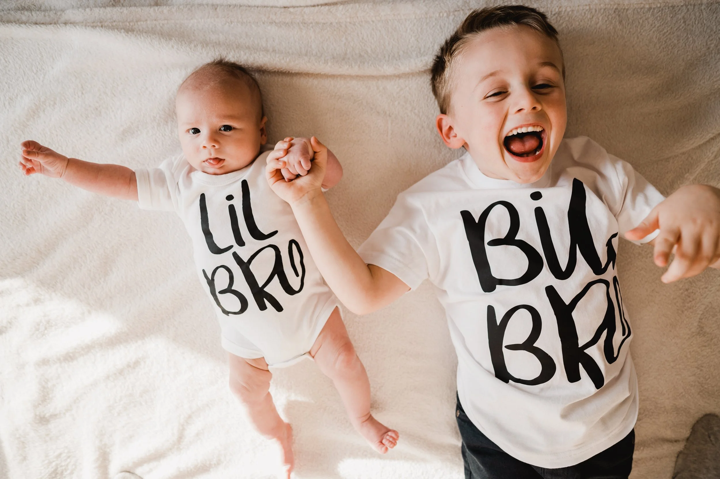 A toddler and a baby lying on a white blanket, dressed in matching white T-shirts with black text, holding hands, with the toddler laughing with an open mouth.